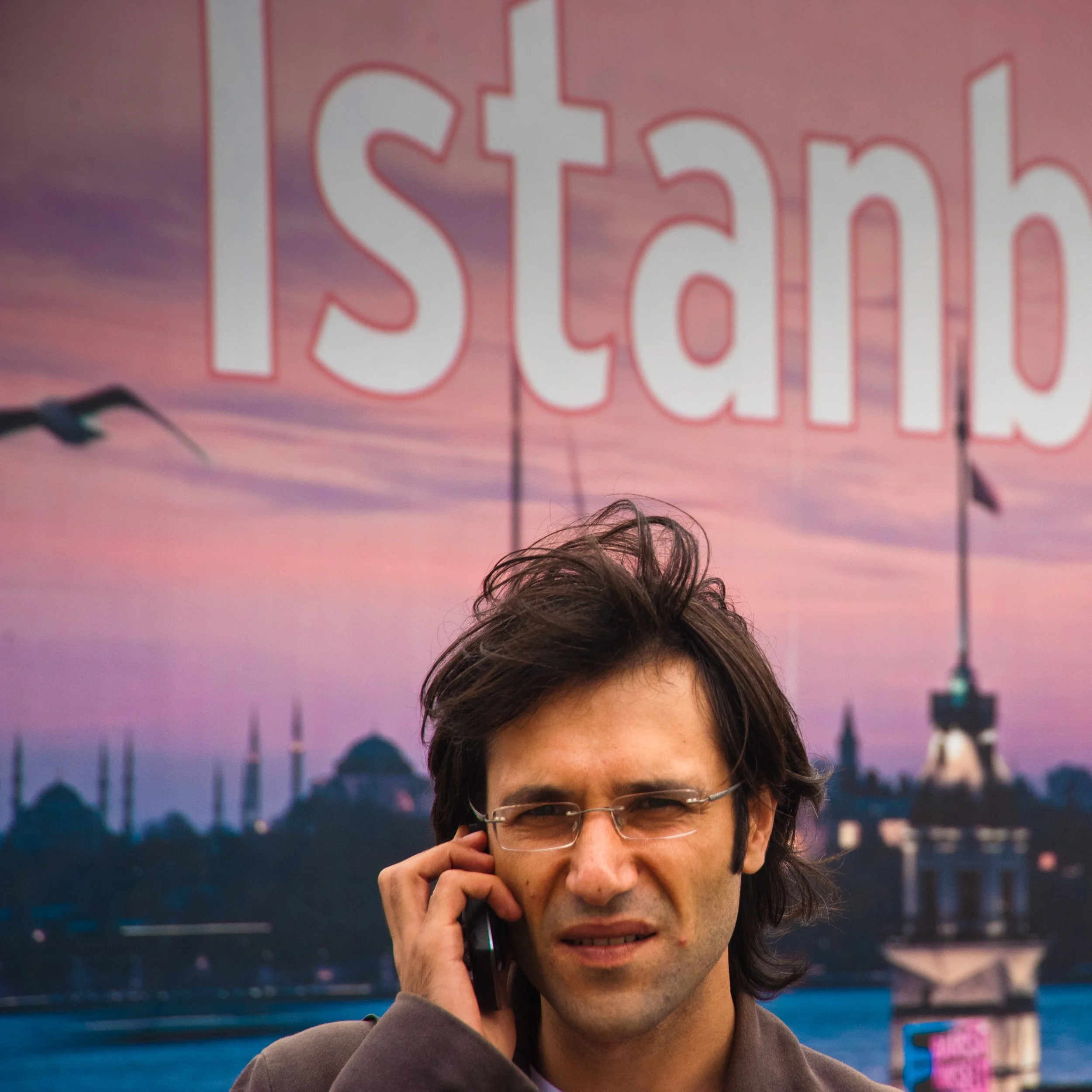A man with dark hair, glasses, and a slight expression of concern is talking on a cellphone in front of a large 'Istanbul' sign. The background shows sunset and landmarks of Istanbul.