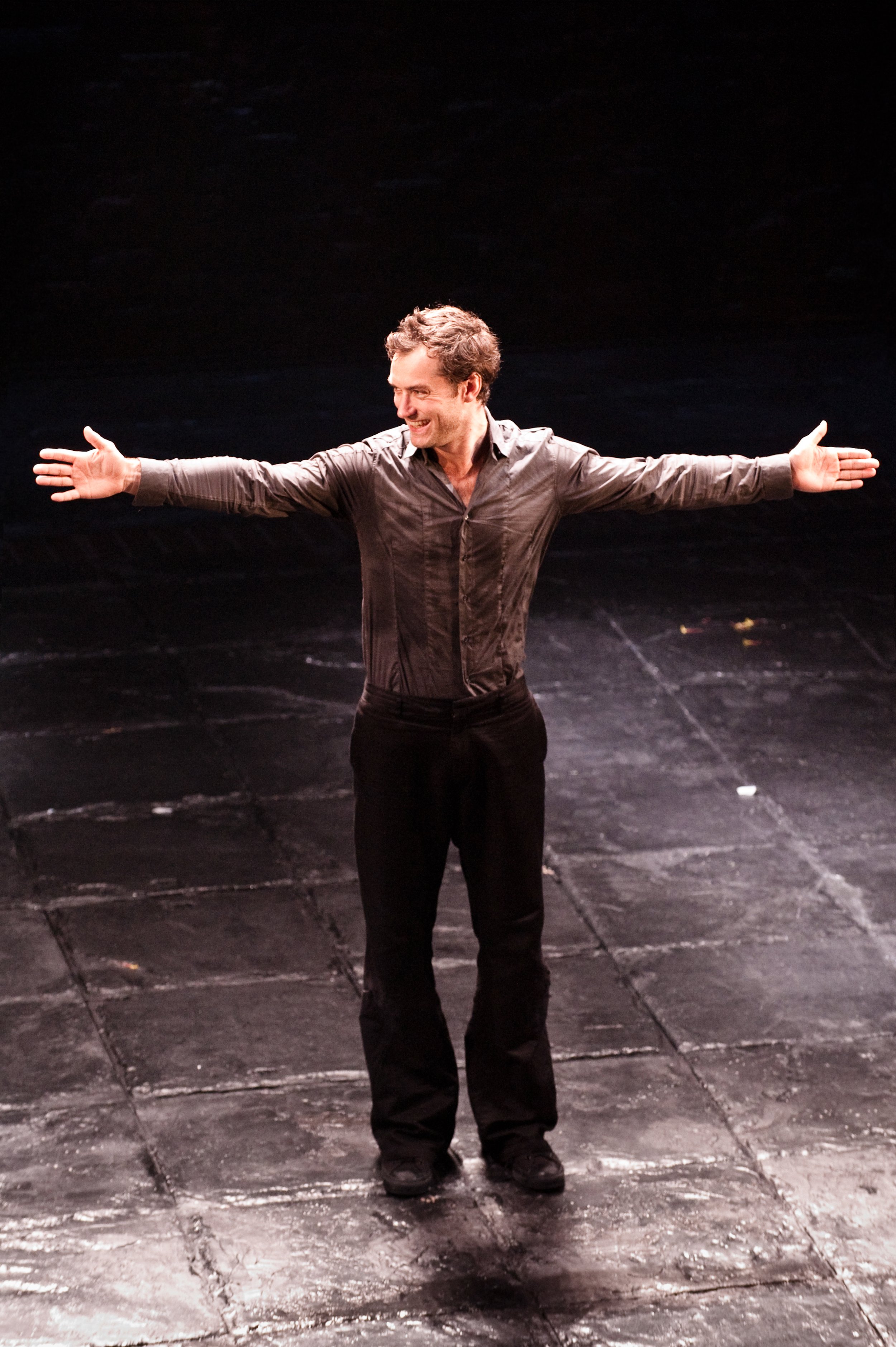 A man standing on a stage with arms wide open, smiling, wearing a gray shirt and black pants.