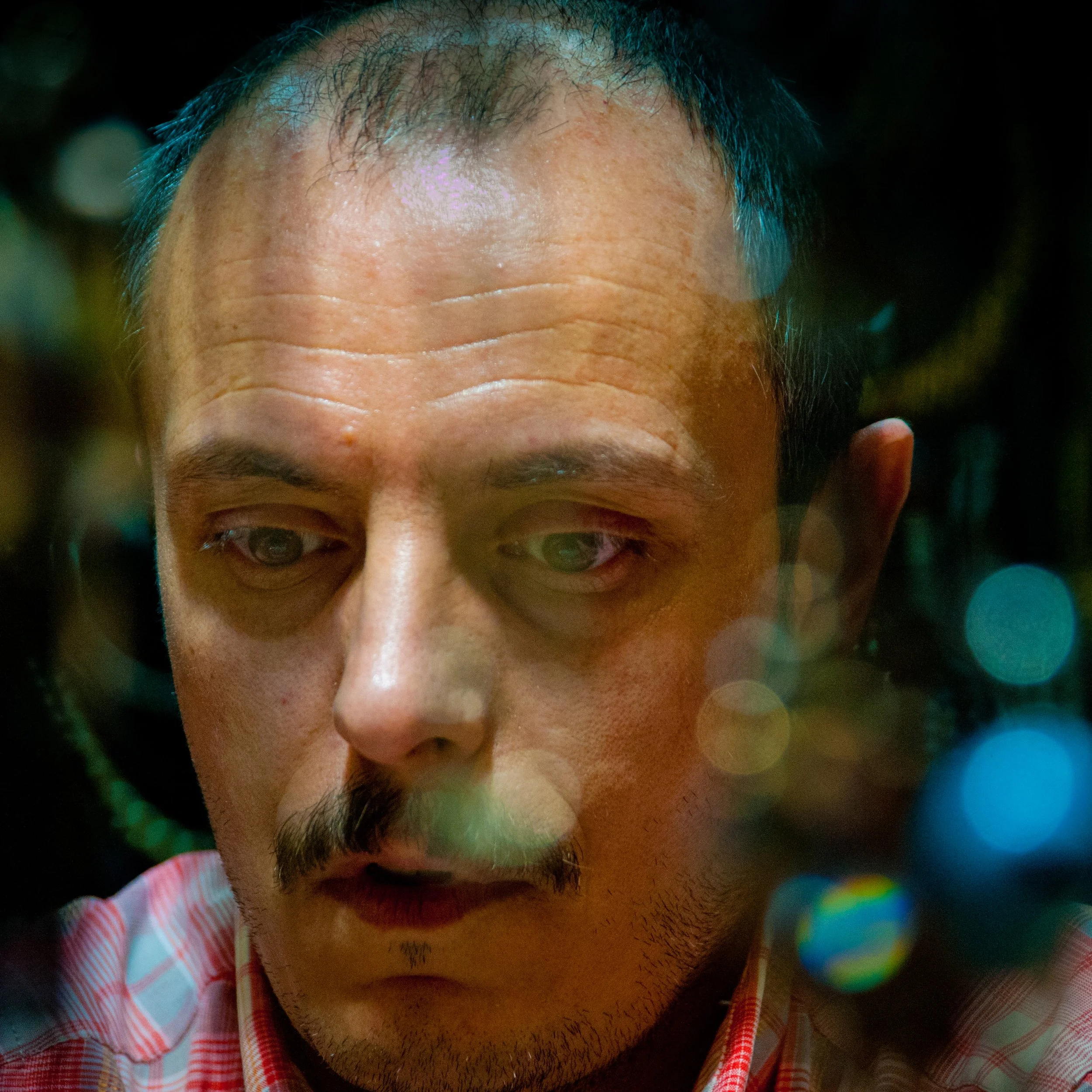 Close-up of a man with short dark hair and a mustache, looking down with a serious expression, illuminated by colorful bokeh lights in the background.
