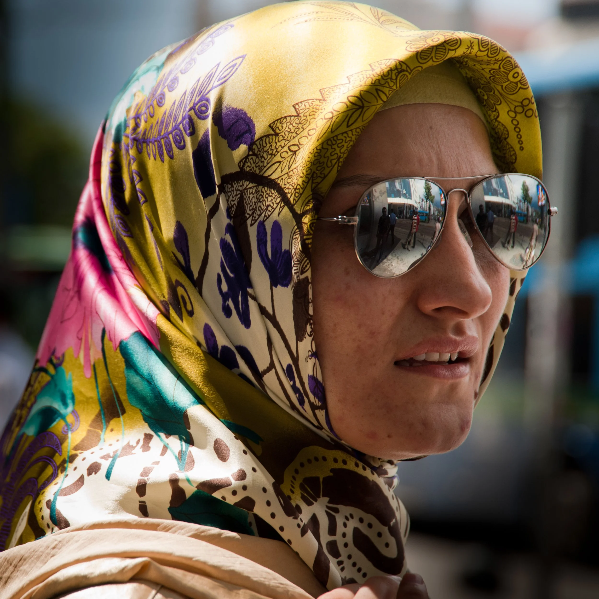 Close-up of a woman wearing a colorful scarf, sunglasses reflective of a city street scene, and a neutral facial expression.