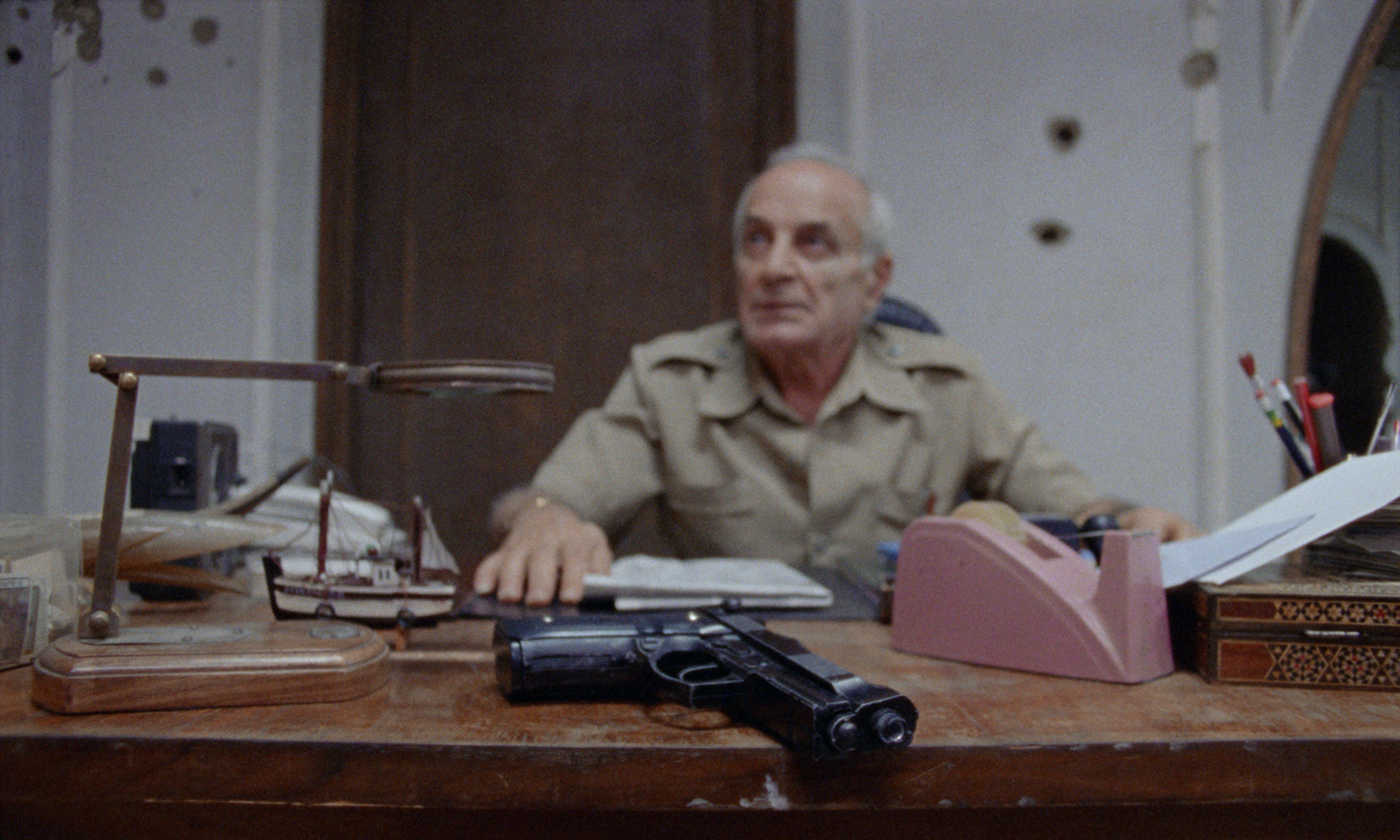 An older man in a beige military uniform sitting at a cluttered desk with a gun, a model ship, papers, and office supplies, looking to his left.