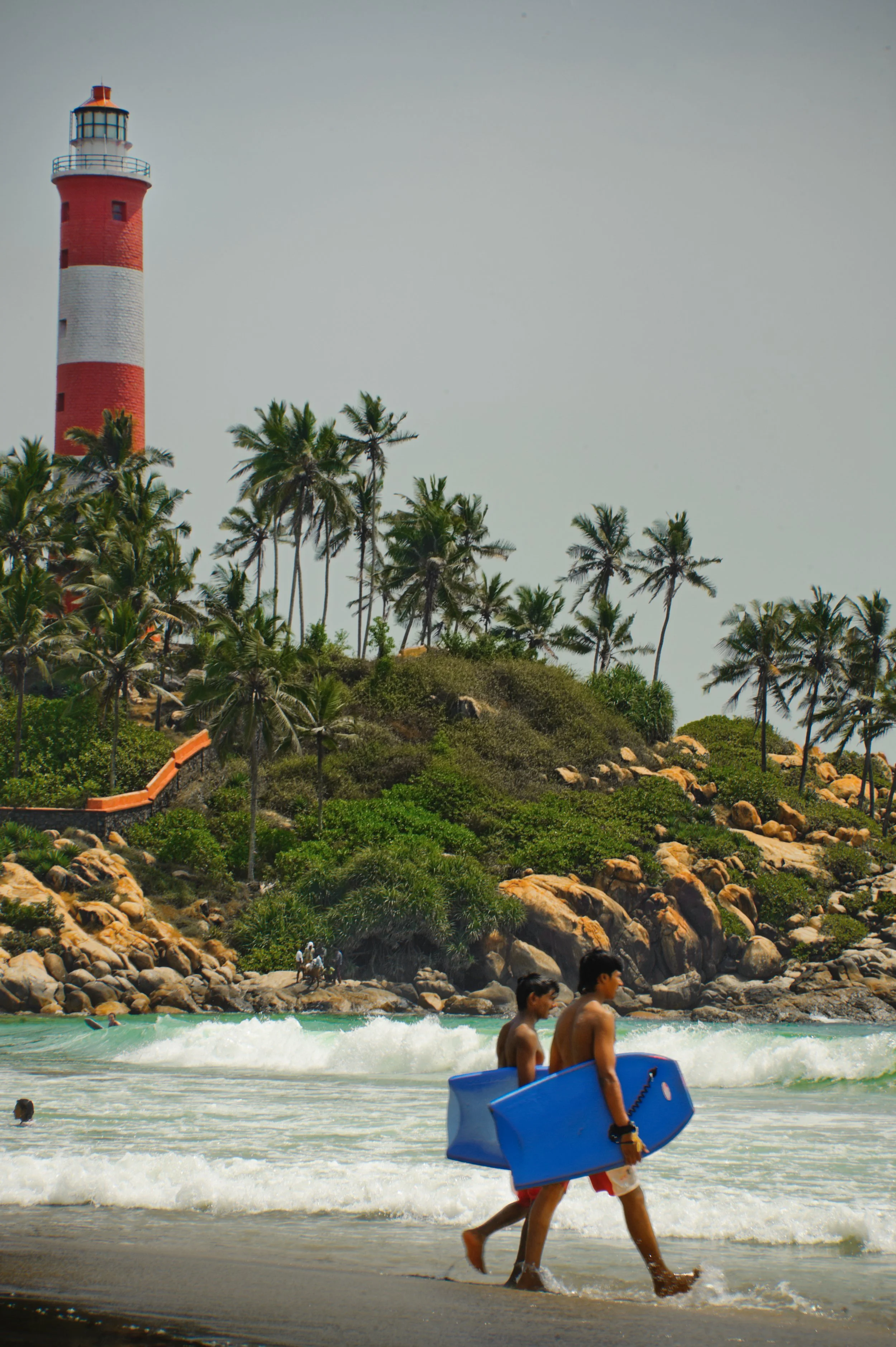 Two boys carrying blue bodyboards walk along the beach shoreline with waves crashing around them. In the background, there is a hill covered with palm trees and rocks, with a red and white striped lighthouse at the top.