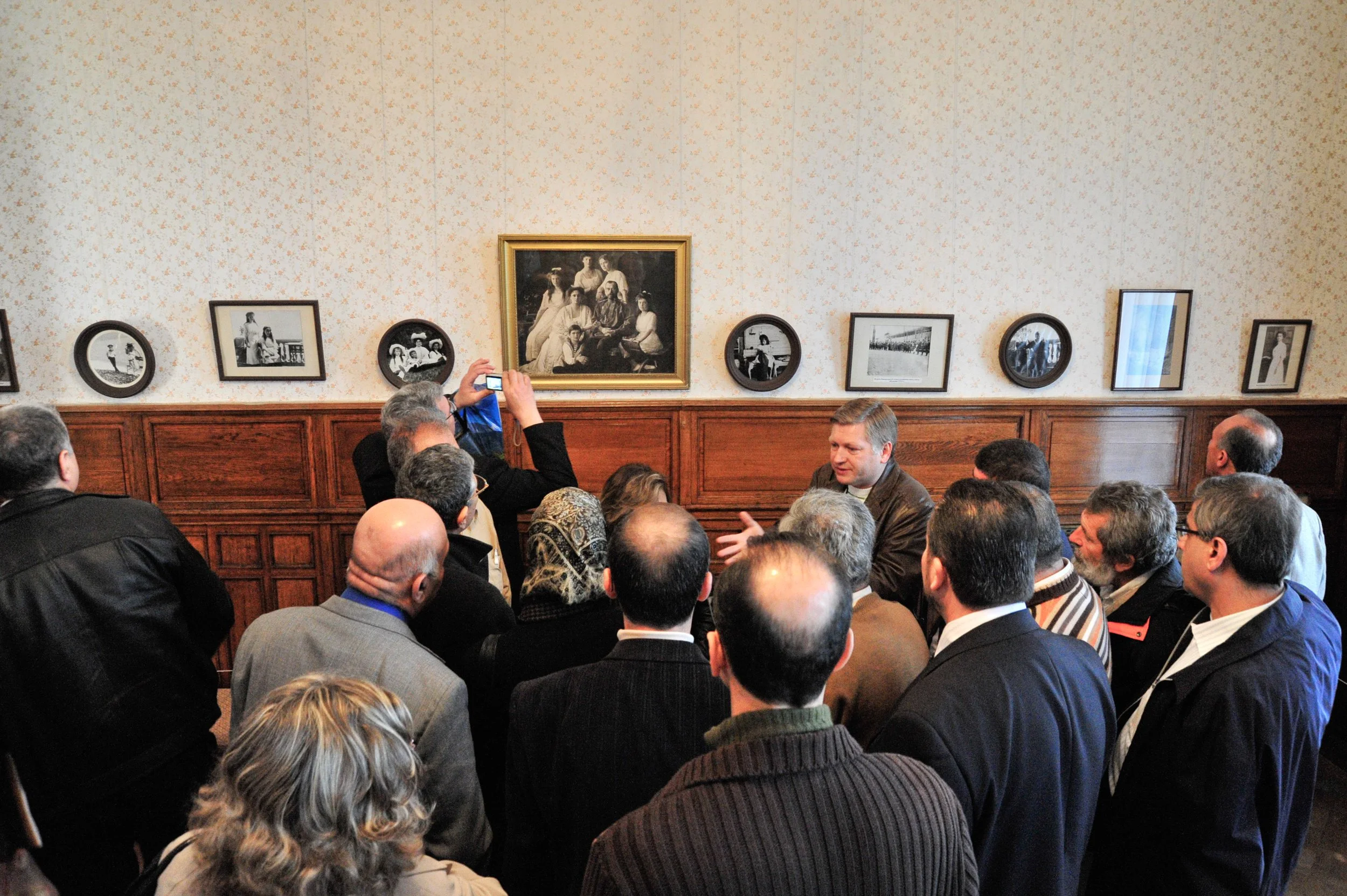 A group of people gathered in a room with wood-paneled walls and vintage photographs, listening to a man speaking, while one person takes a photo of the scene.
