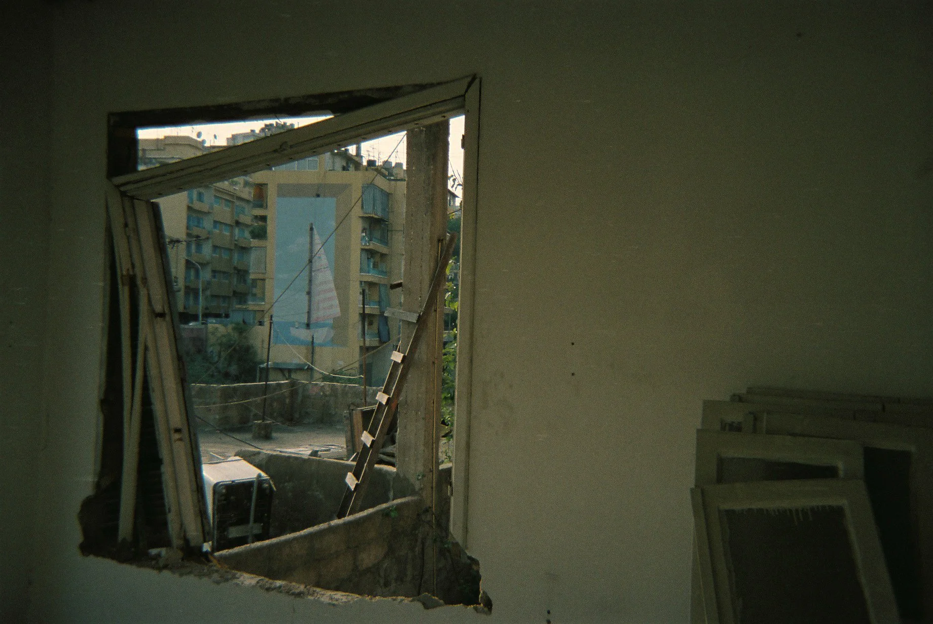 View from a damaged building window showing a cityscape with multiple apartment buildings and laundry hanging outside, with some construction materials or window frames stacked inside.