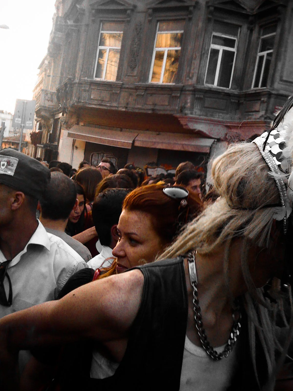 People gathered in a crowded street surrounded by old buildings with ornate architecture in the background, some wearing fashionable clothing and accessories.
