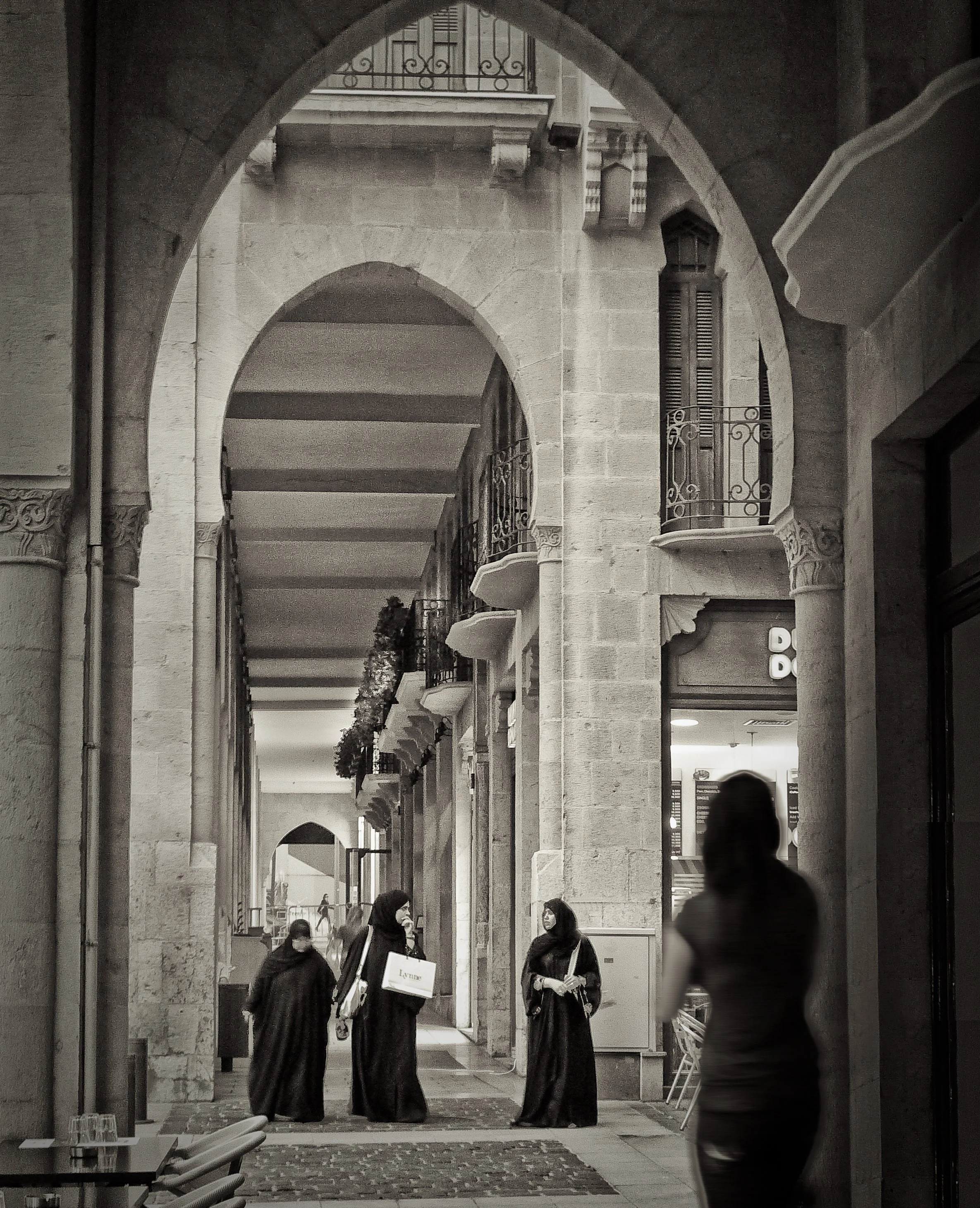 A black and white photo of a covered walkway in an urban area with archways, balconies with decorative railings, and people walking and conversing. There are three women in traditional attire holding shopping bags and a woman in casual clothing walki