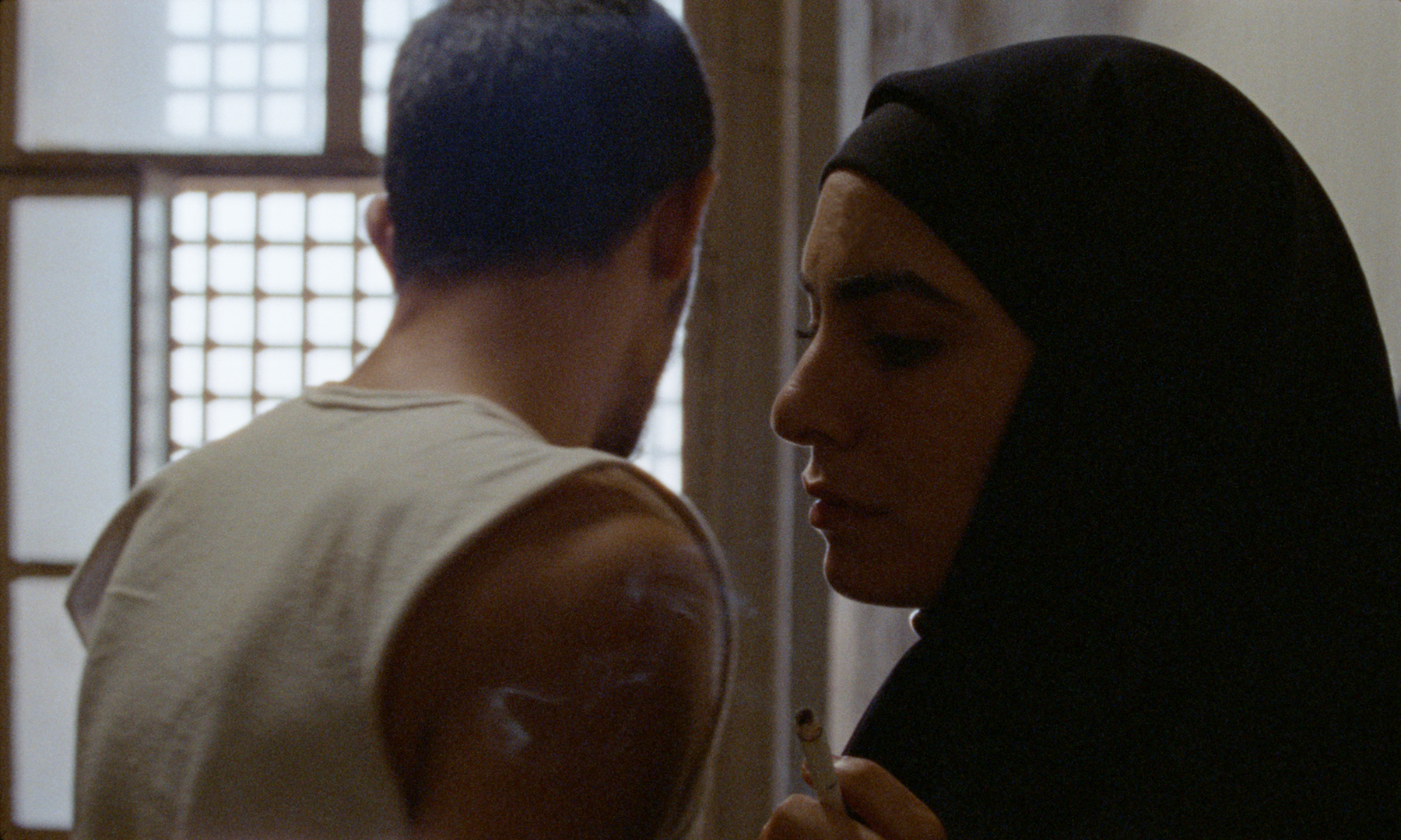 A man and a woman face each other close, with the woman looking down and the man looking at her neck, holding a small object near her neck. They are indoors with patterned windows in the background.