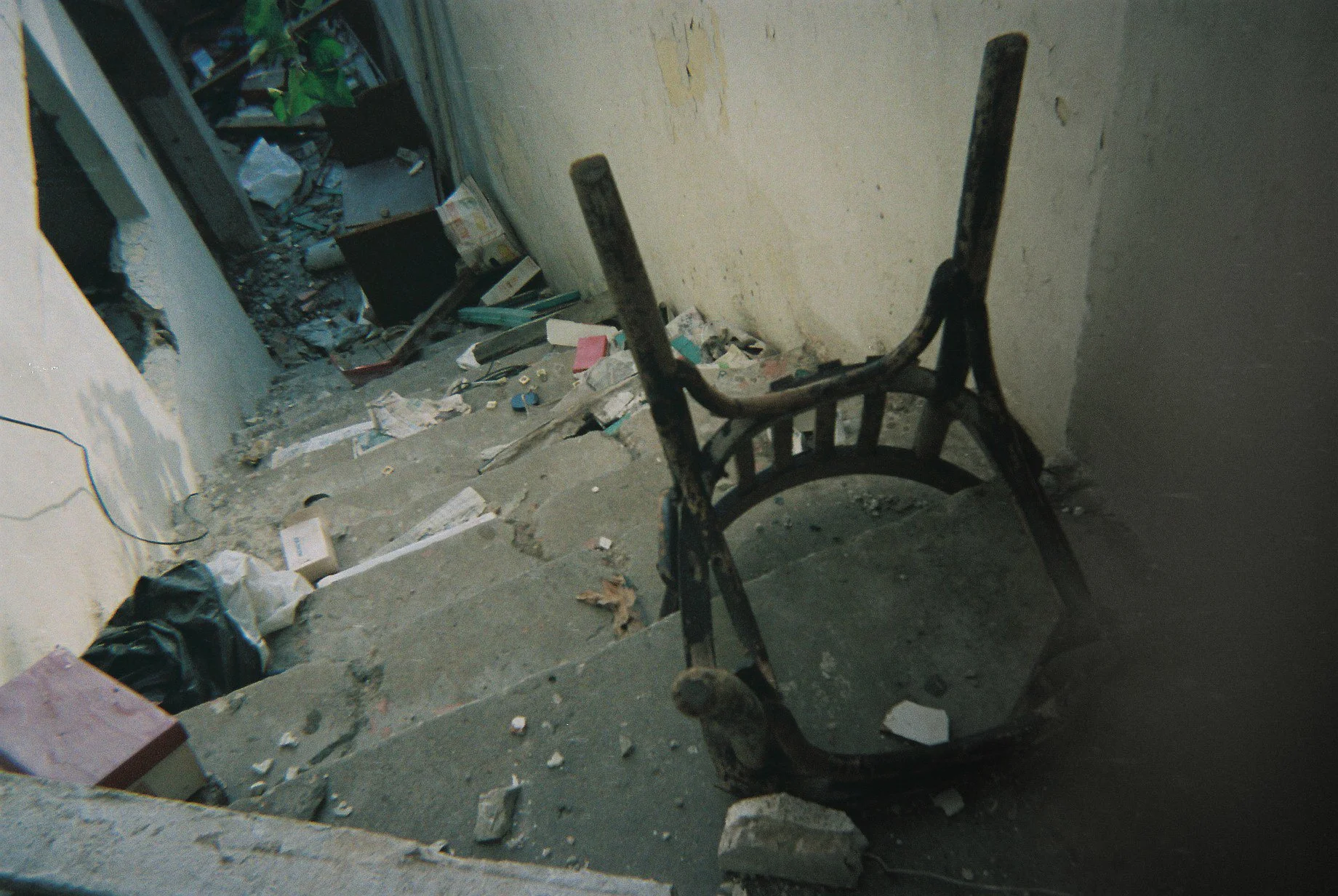 A decrepit room with a broken chair lying on the concrete floor surrounded by trash and debris. The wall is damaged and cluttered with disorganized items visible in the background.