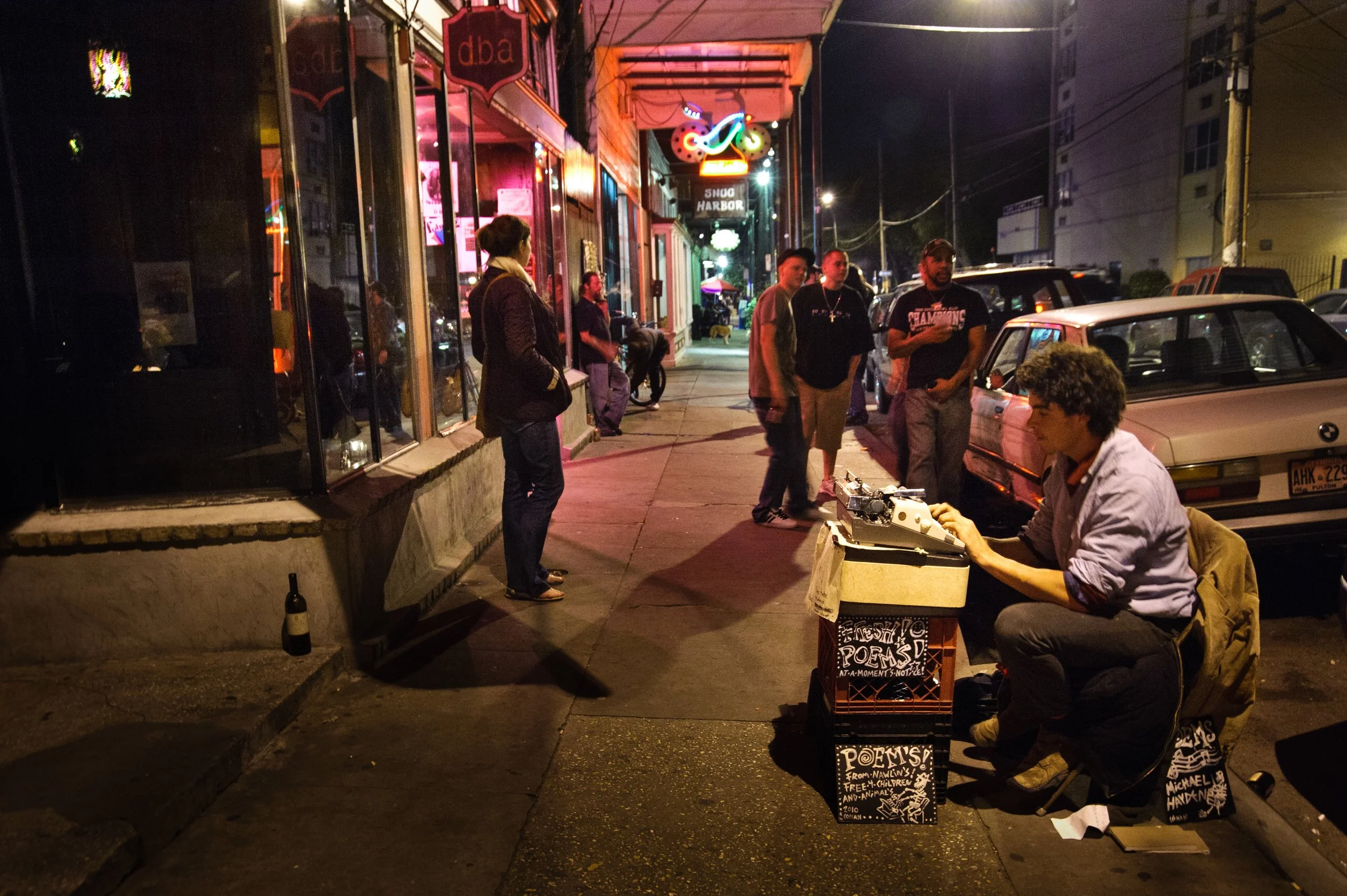 A street scene at night with people gathered around a man performing poetry at a small outdoor stand, decorated with signs reading 'Poems!'. The performer is sitting on a folding chair, working on a typewriter or similar device. Several cars are park