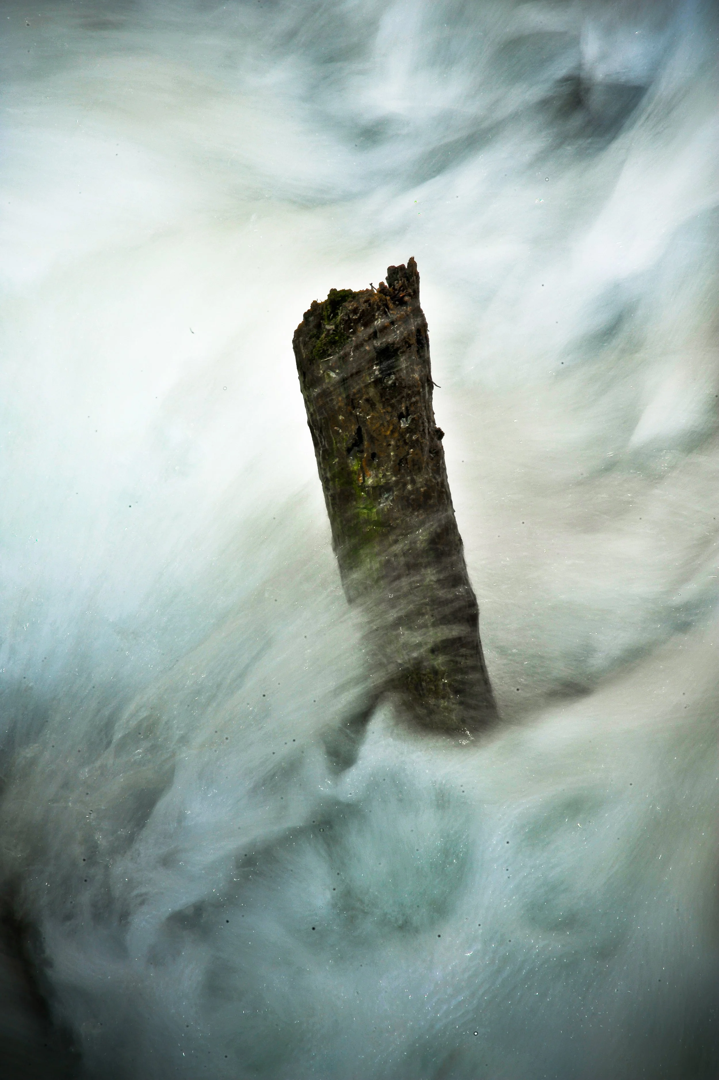 Close-up of a weathered wooden post surrounded by fast-moving water, creating a misty and turbulent scene.