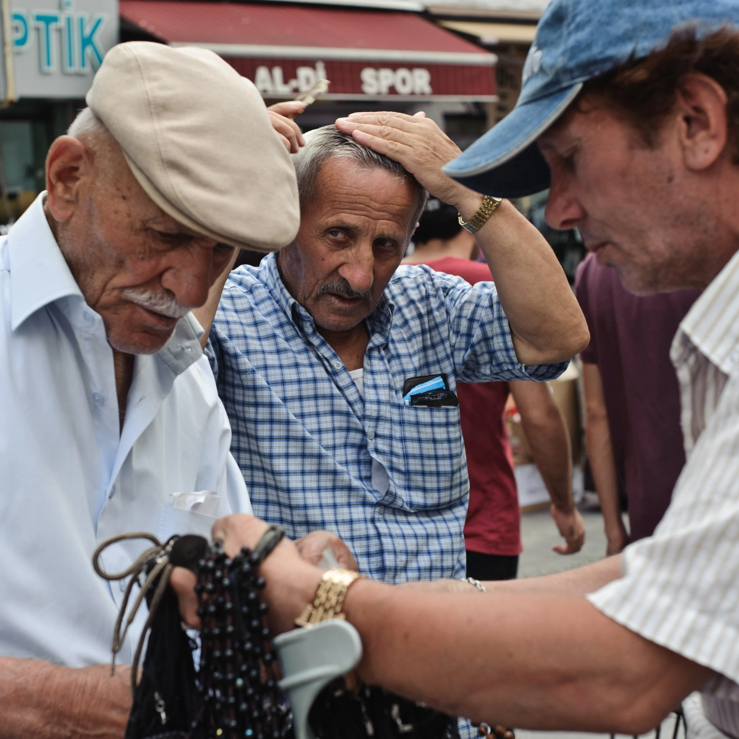 Three men are closely examining a wristwatch in a busy outdoor market, with buildings and a sign in the background.