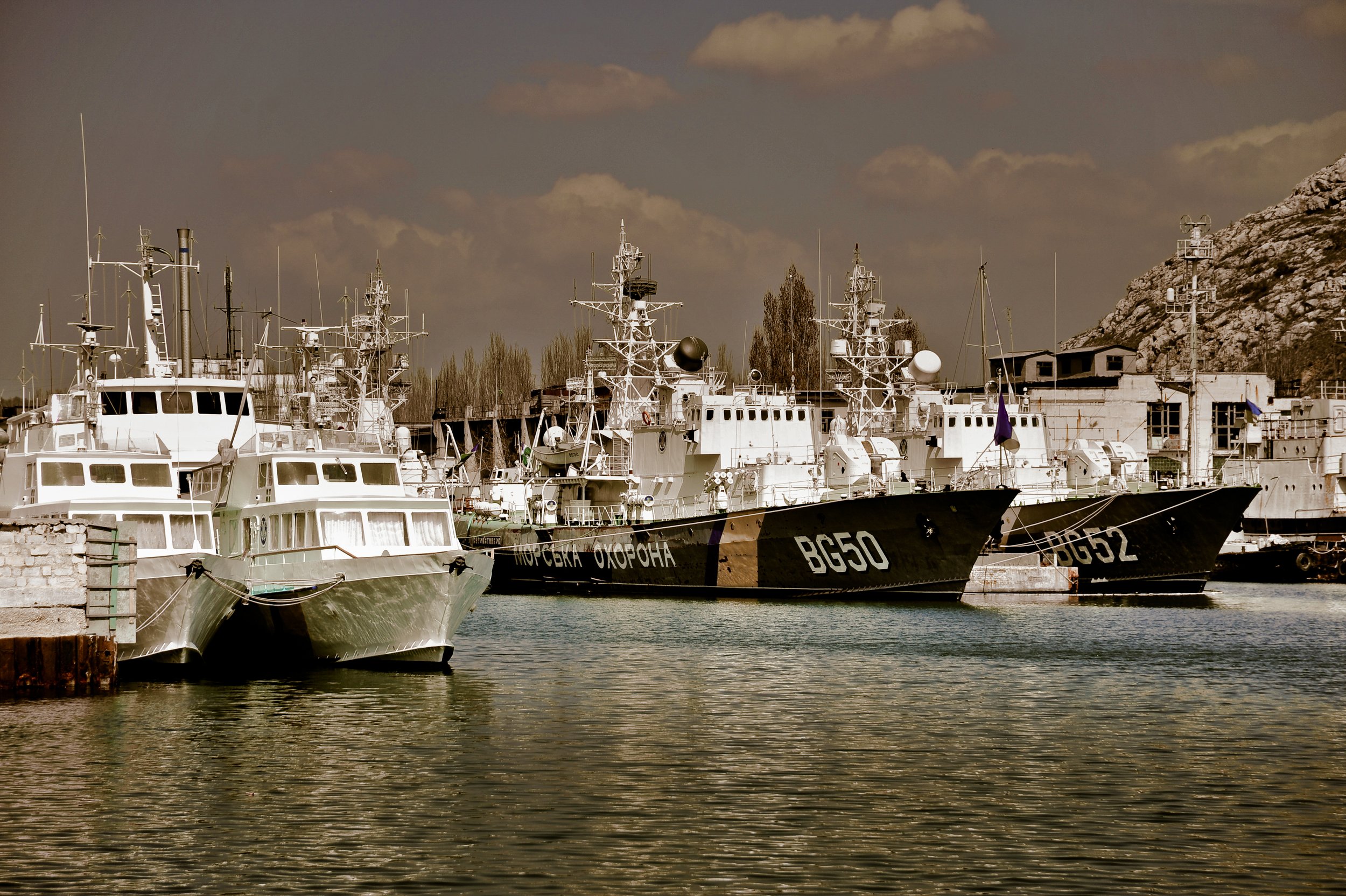 Several military ships docked at a harbor with rocky hills in the background.