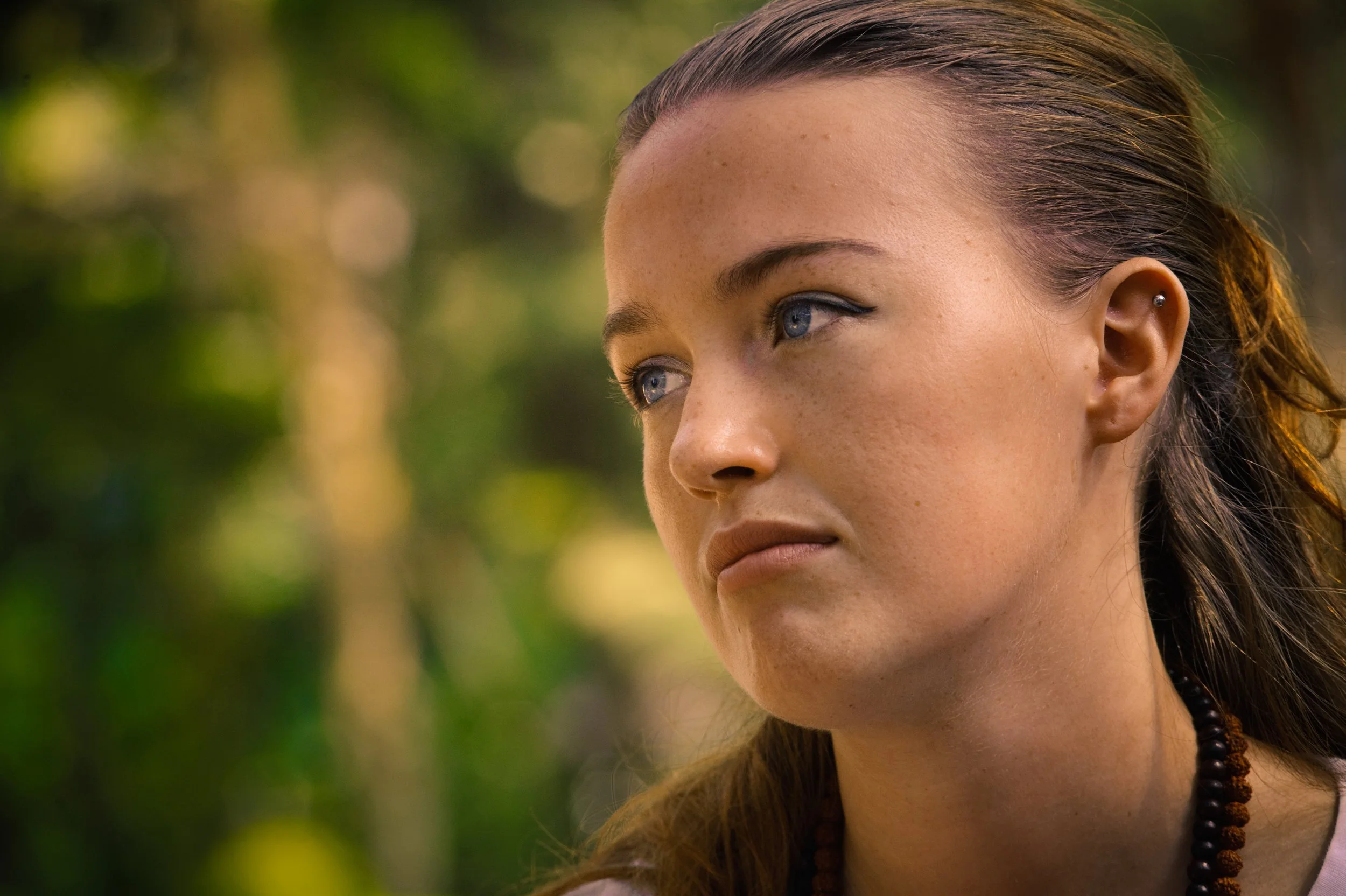 Close-up of a young woman with blue eyes and freckles, looking thoughtfully into the distance, outdoors with blurred green foliage in the background.