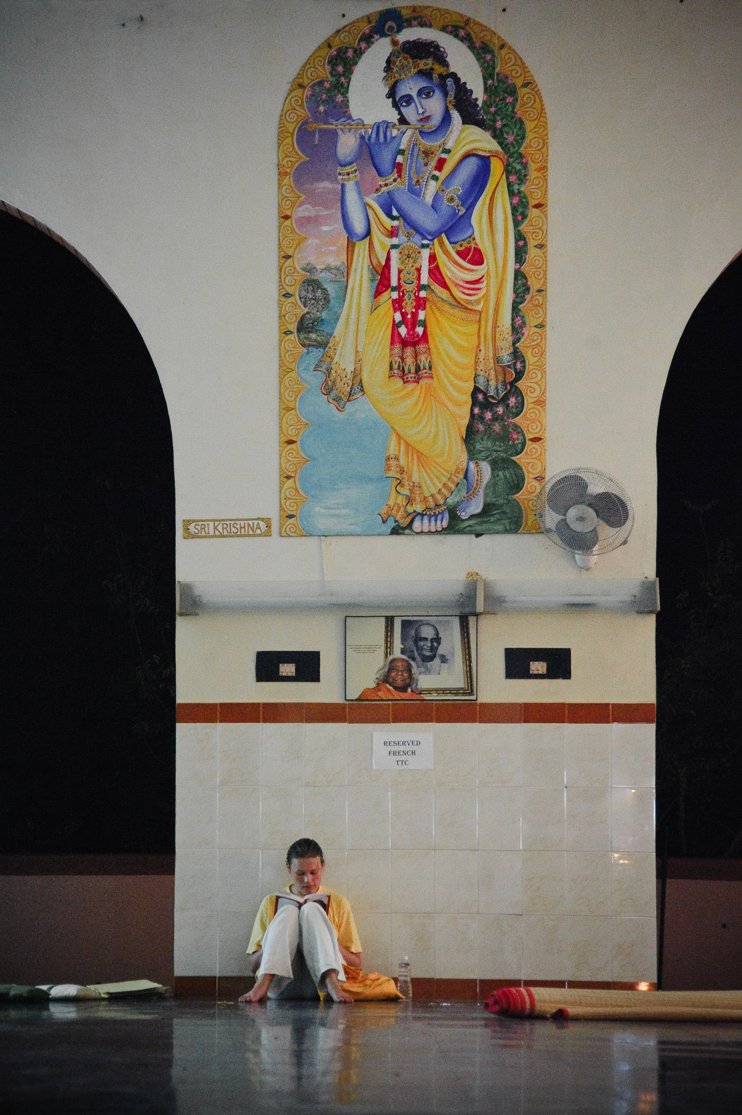 A girl sitting on the floor reading a book in a temple with a large painting of Lord Krishna playing a flute hanging on the wall above her. There is a small framed photo of a saint or spiritual leader and a sign that reads 'Reserved French TTC' in fr