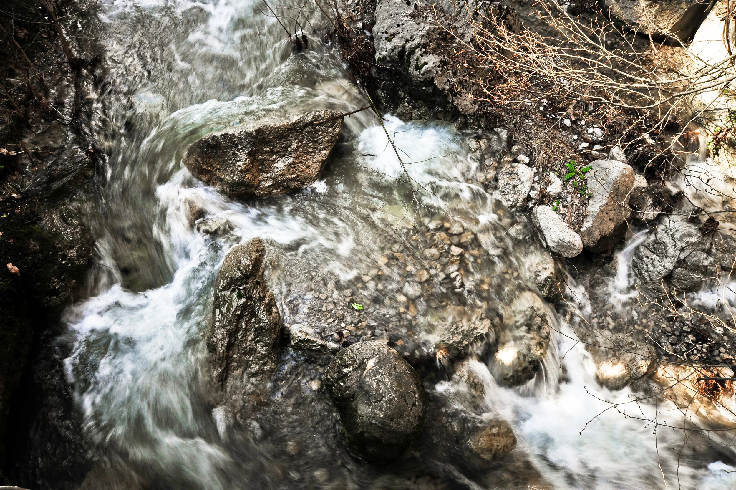 A rocky stream with moving water flowing over and around large rocks and boulders, surrounded by small branches and sparse vegetation.