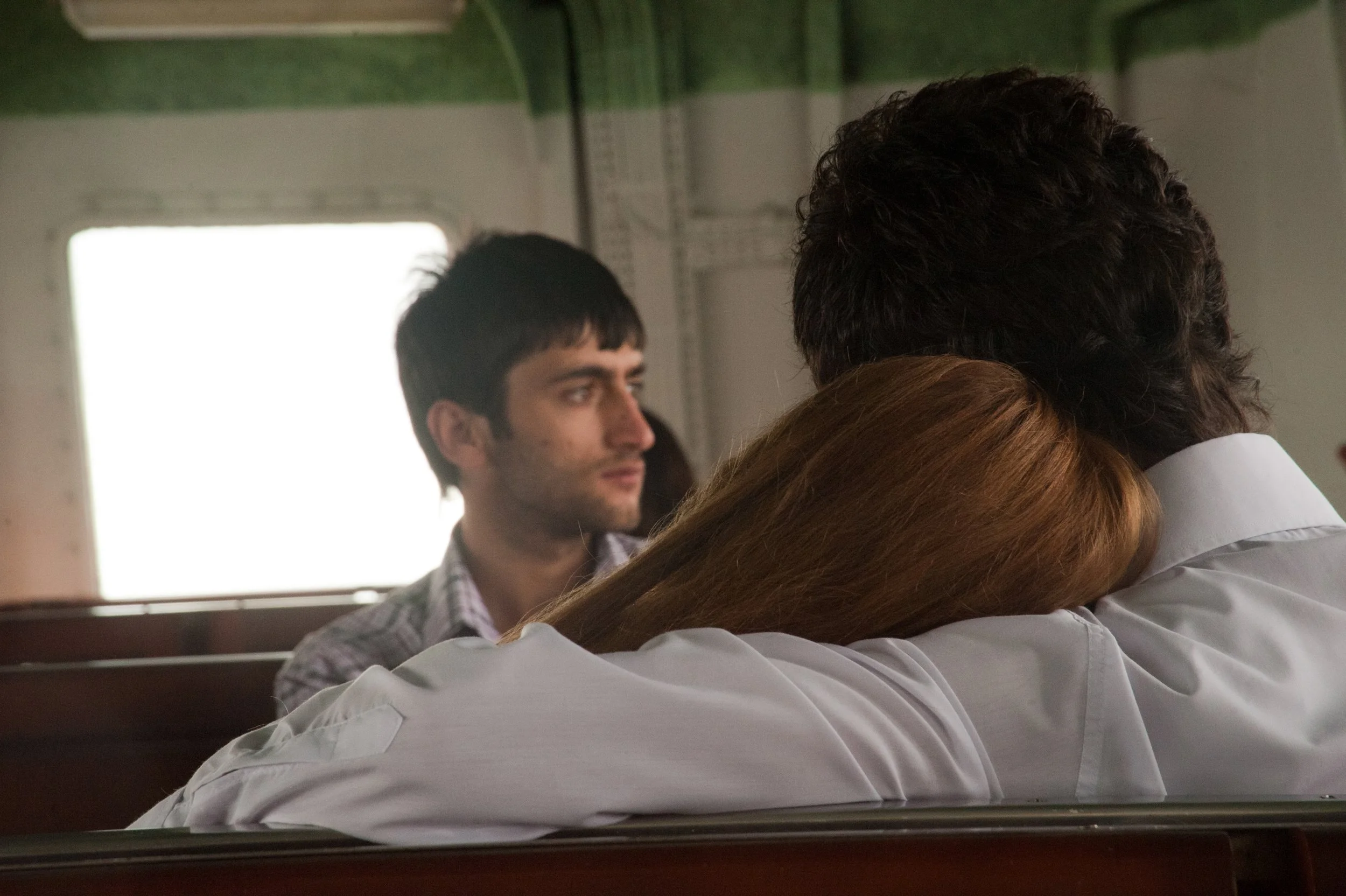 A woman with red hair resting her head on a man's shoulder while sitting on a train, with a young man appearing deep in thought in the background.