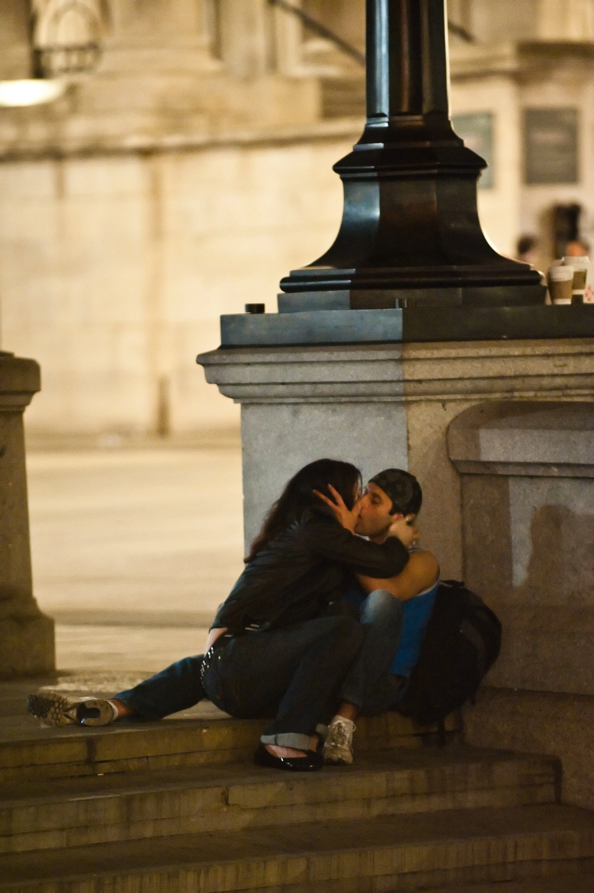 A couple kissing on the steps under a lamp post at night, near a stone building.