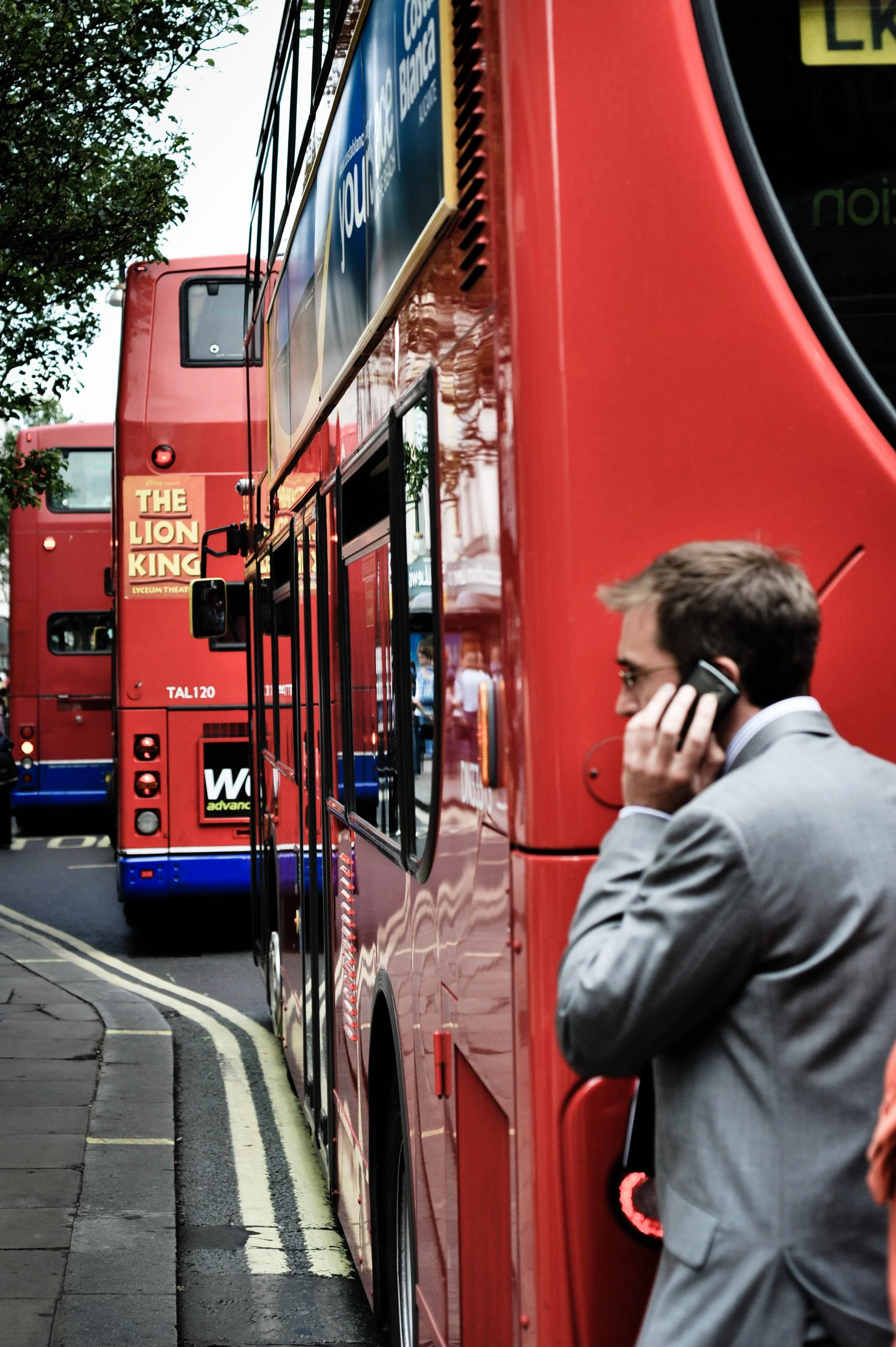A man in a gray suit is talking on a cellphone while standing next to a red double-decker bus on a city street. The bus has a sign advertising 'The Lion King' musical. Another red double-decker bus is visible behind it.