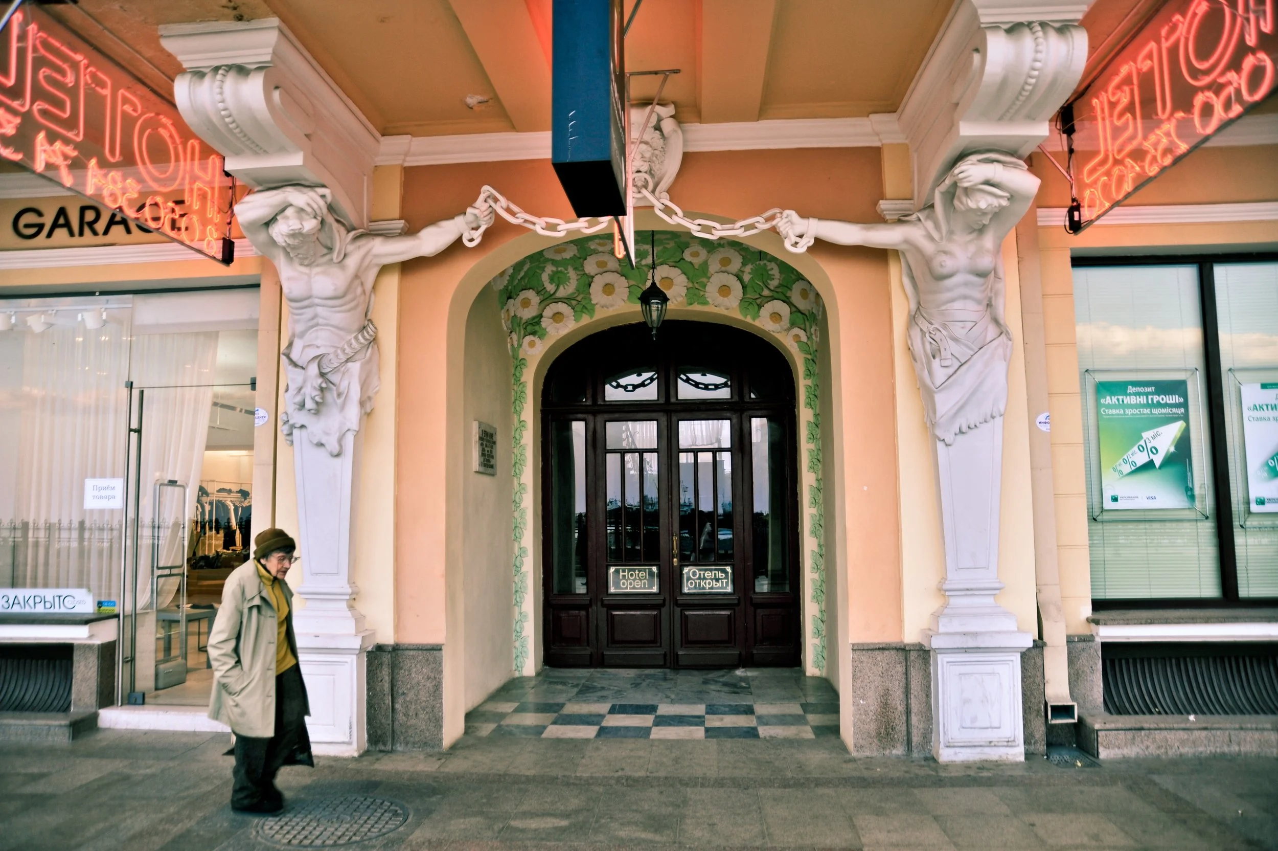 Front entrance of a building with two white sculpted figures supporting a structure above the doorway. The figures are muscular and women with draped clothing, holding chains. The doorway has glass doors with signs indicating the hotel is open, and a