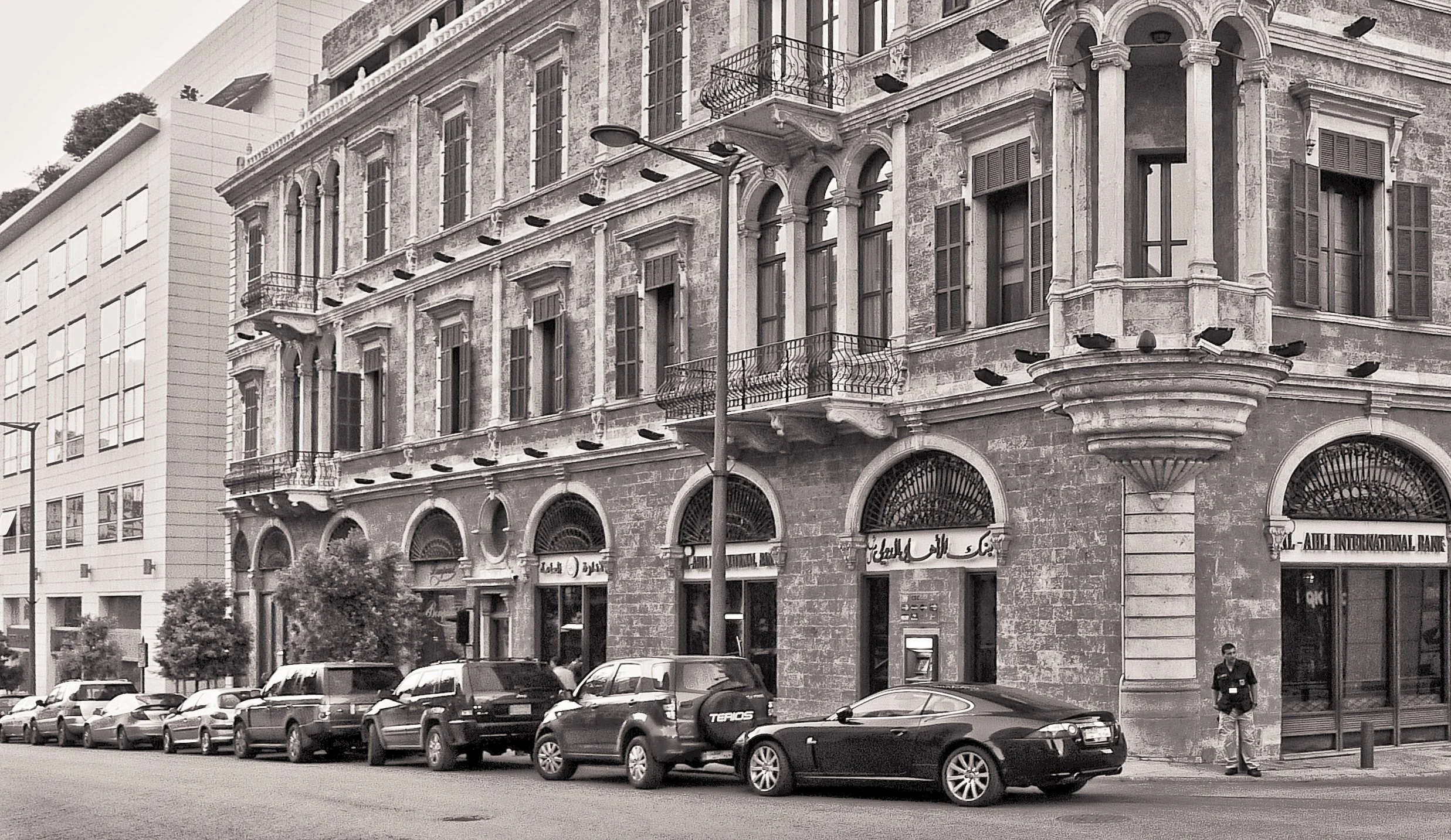 Street view with parked cars in front of an old, multi-story building with decorative balconies and arched windows, alongside a modern building to the left. A man stands near a bank branch entrance.