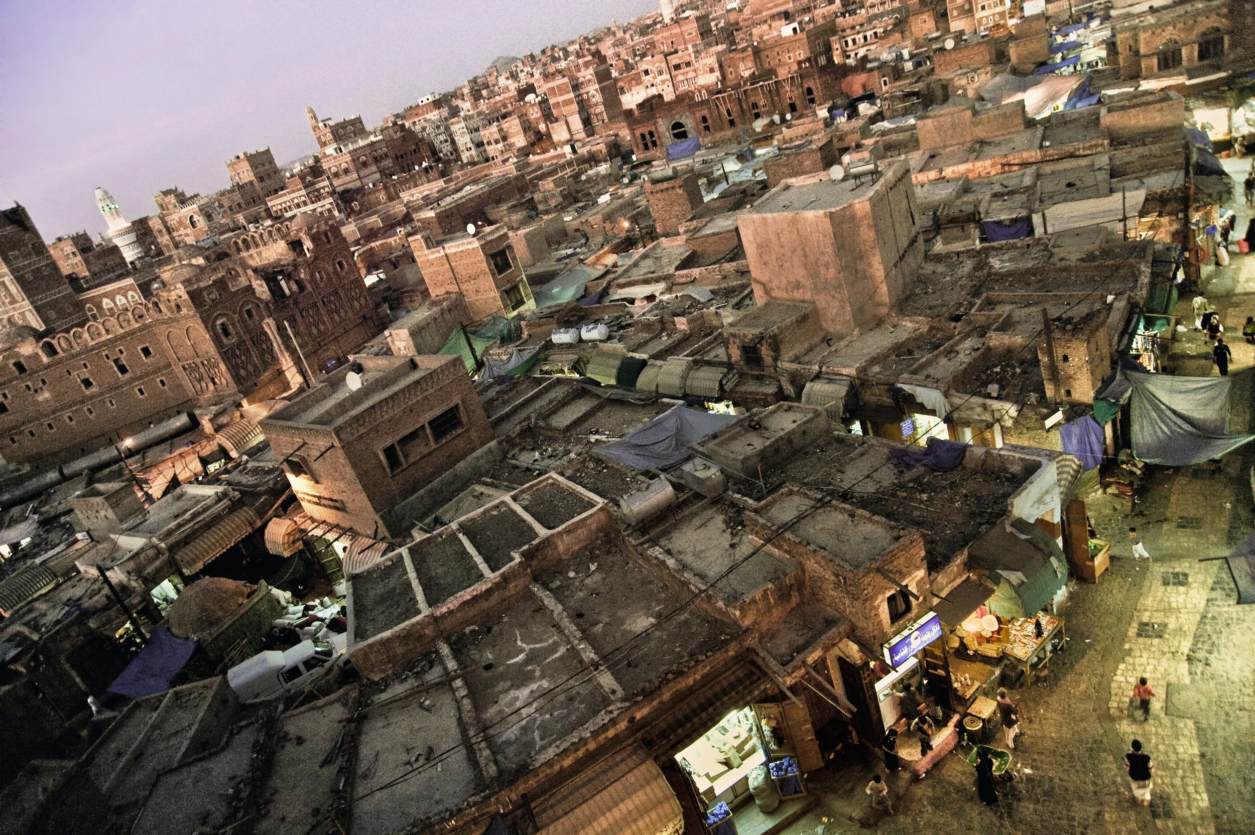 Aerial view of a crowded marketplace in a Middle Eastern or North African city with closely packed buildings and small shops, some with brightly lit signs, during dusk.