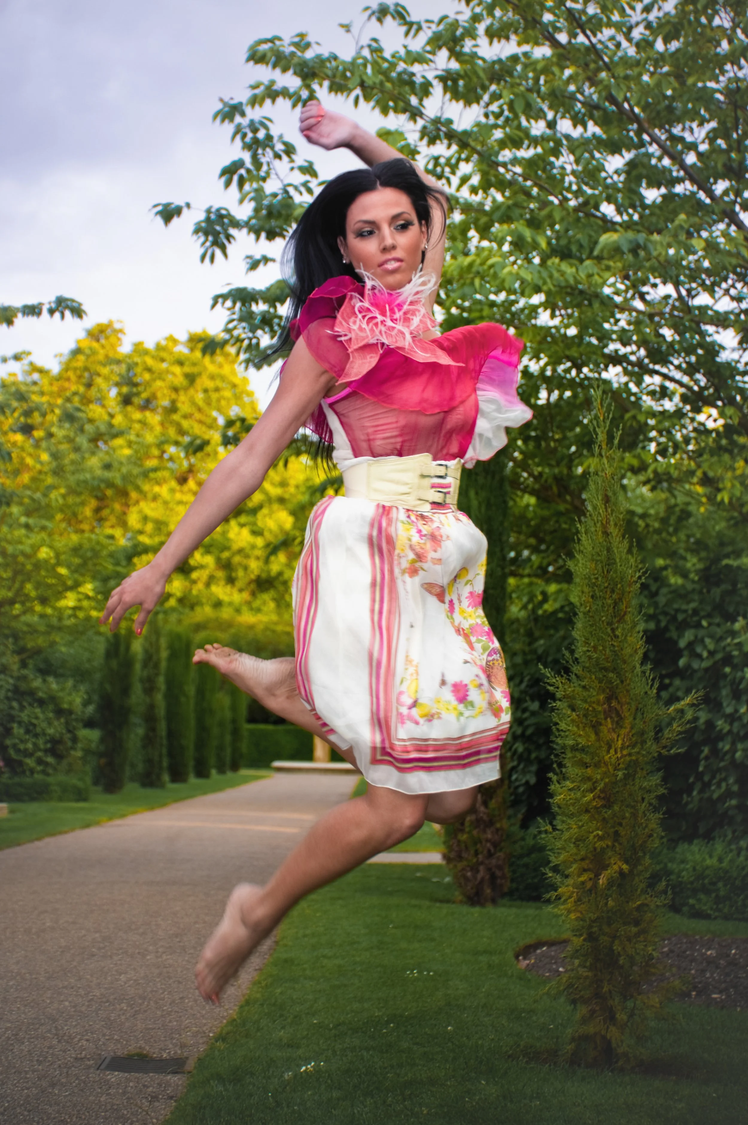 A woman with long black hair jumping outdoors in a garden, wearing a colorful dress with a pink and white top and a floral skirt.