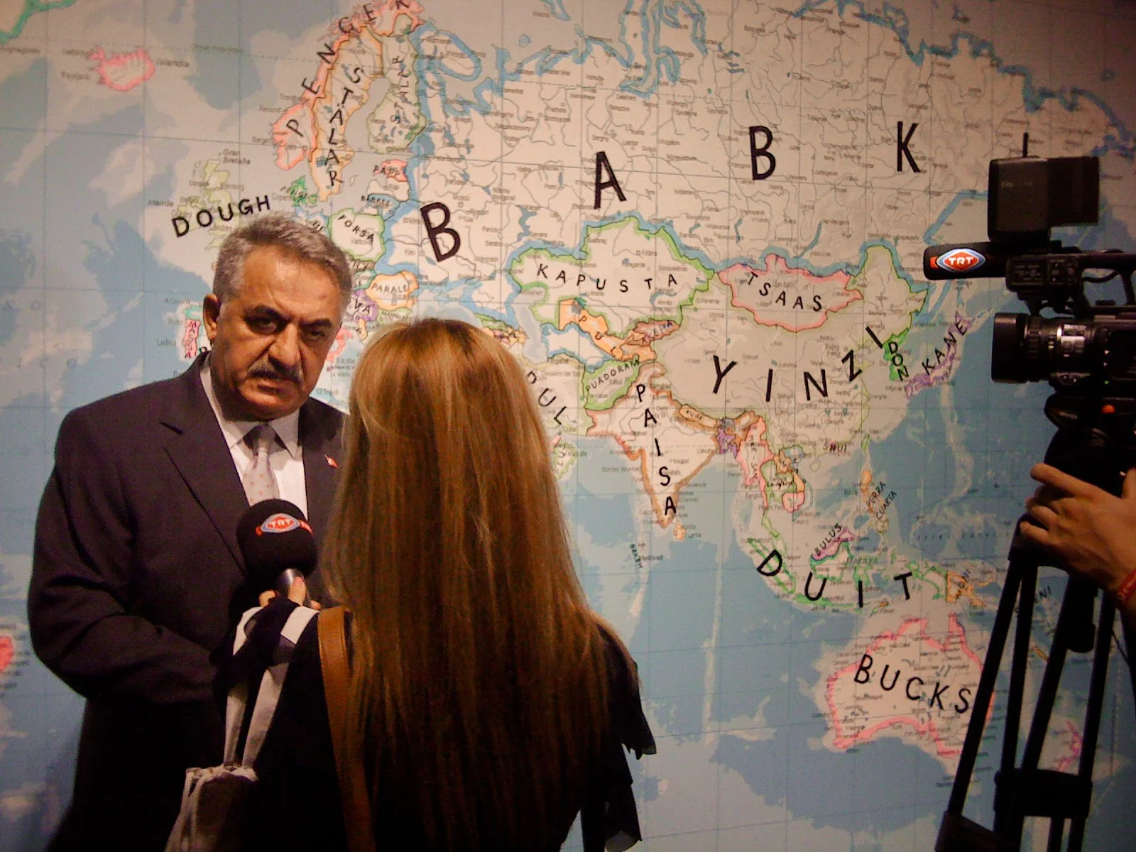A man in a suit being interviewed by a woman in front of a world map featuring the continent of Asia with country names written in black and outlined in different colors, with a camera recording the interview.