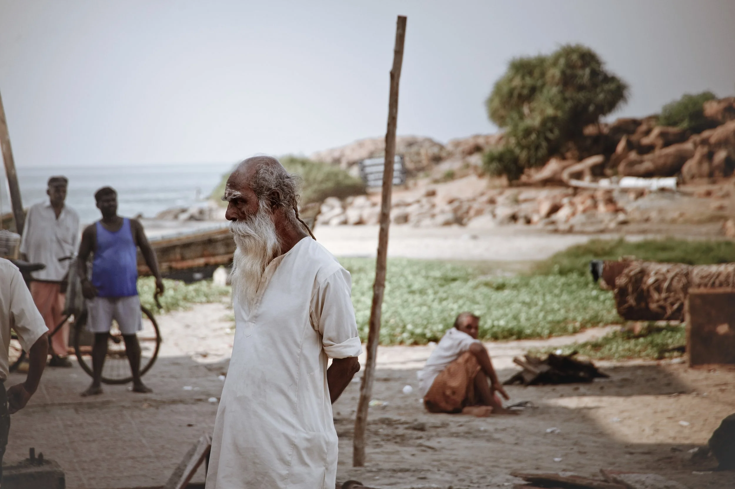 An elderly man with a white beard and traditional white clothing stands in a rural outdoor setting, with a few other men and natural landscape in the background.