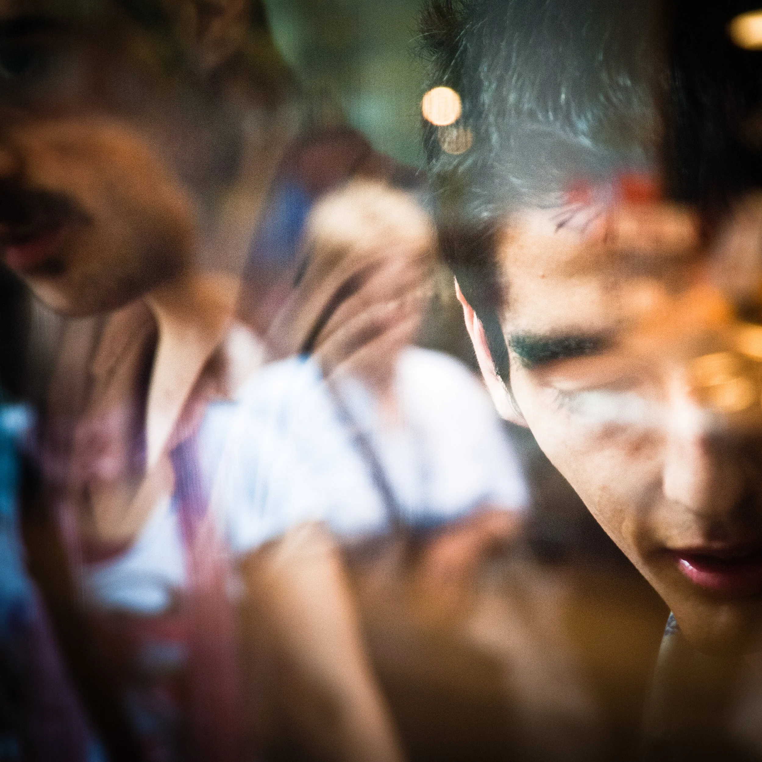 Close-up of a young man with dark hair, smiling and looking down, with blurred people behind him in a lively indoor setting.