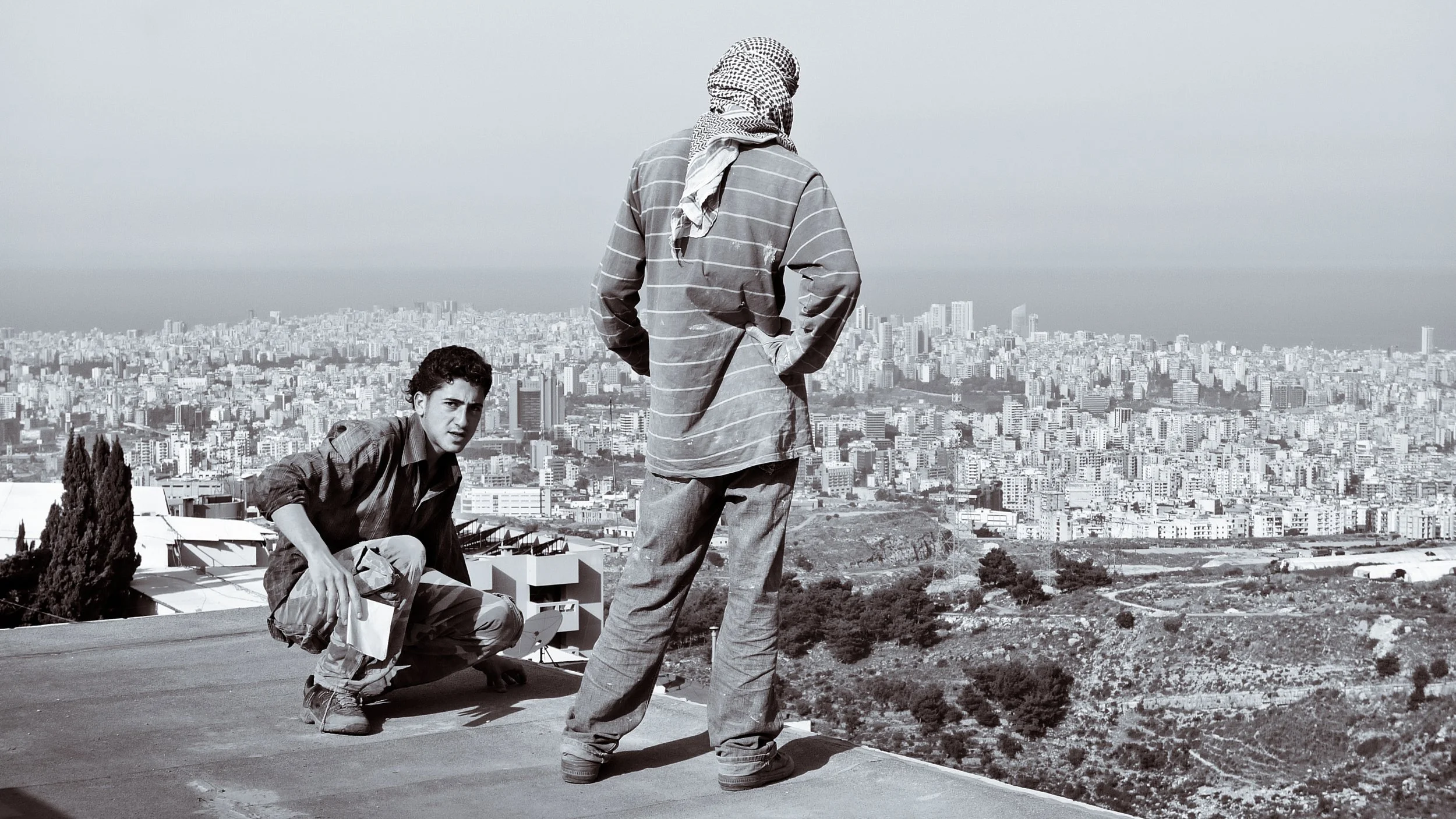Two men on a rooftop overlooking a cityscape in black and white. One man is kneeling on the ground, looking directly at the camera, while the other man stands with his back to the camera, gazing at the city.