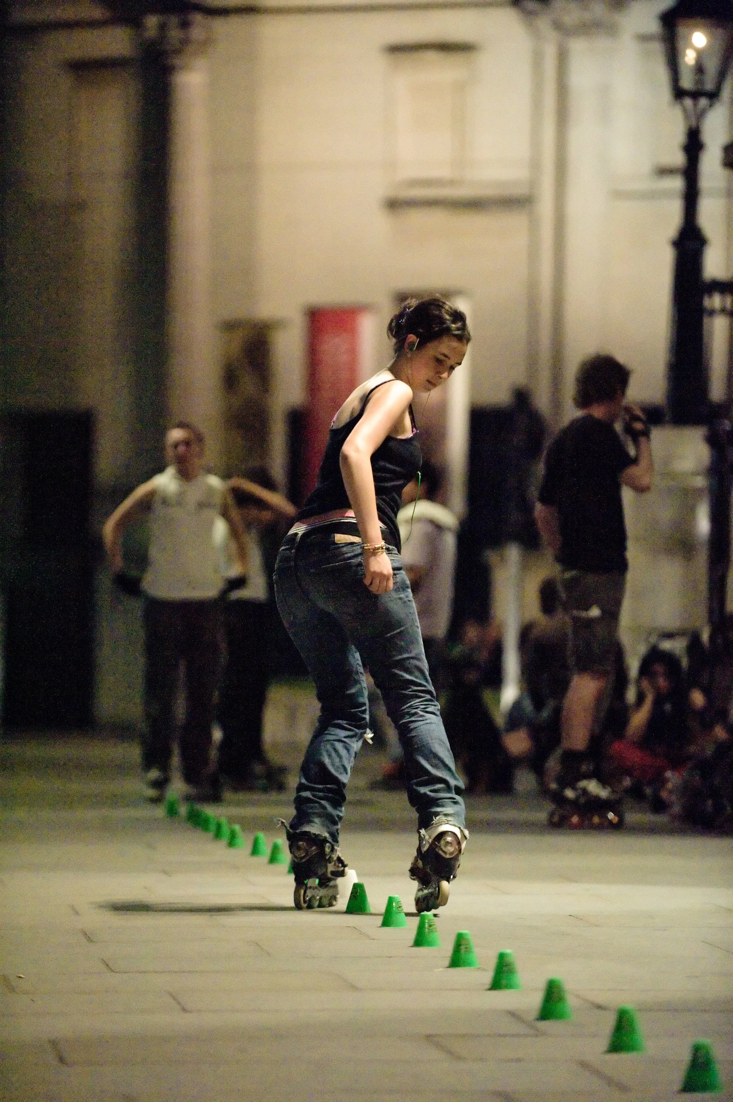 Girl rollerblading through a line of green cones on an outdoor pavement at night.