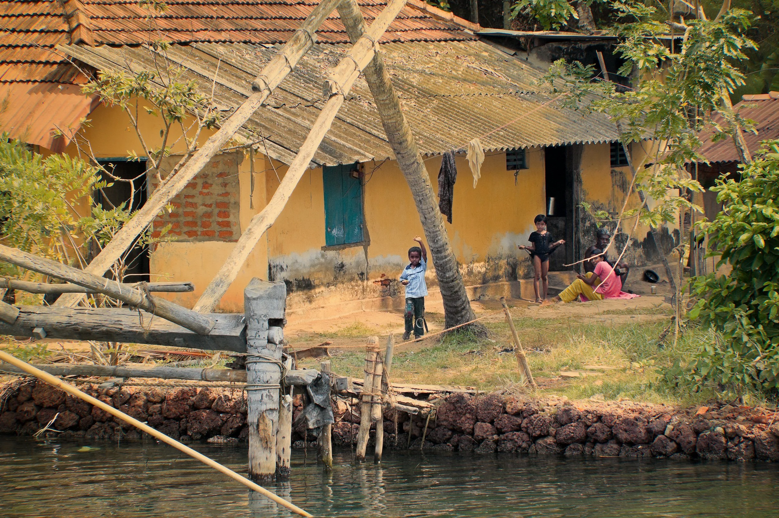 A yellow house with a red tile roof, weathered walls, and a blue door sits by a water body. Four children are playing outside, with one holding a stick, another standing, and two sitting on the ground. There are trees and a fence near the house.