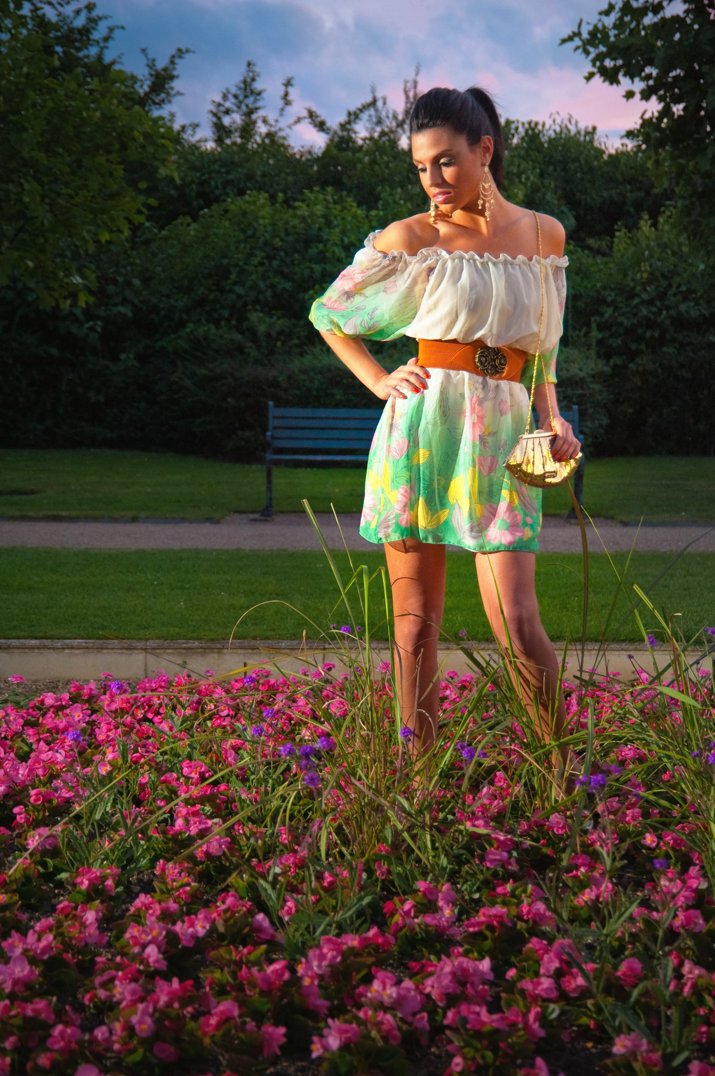 A woman standing in a garden at sunset, wearing a floral off-shoulder dress, holding a small purse, with pink flowers and greenery around her.