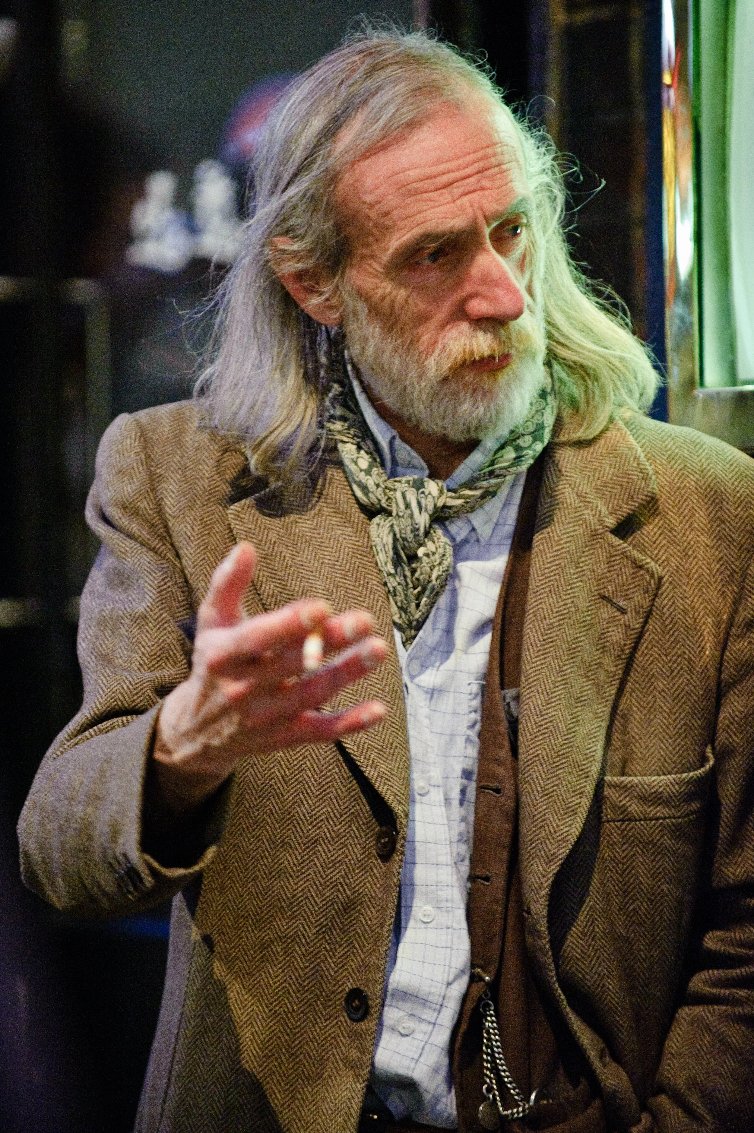An elderly man with long gray hair and a beard, wearing a brown blazer, a patterned scarf, and a checkered shirt, is holding a cigarette and looking out a window in a bar or café.