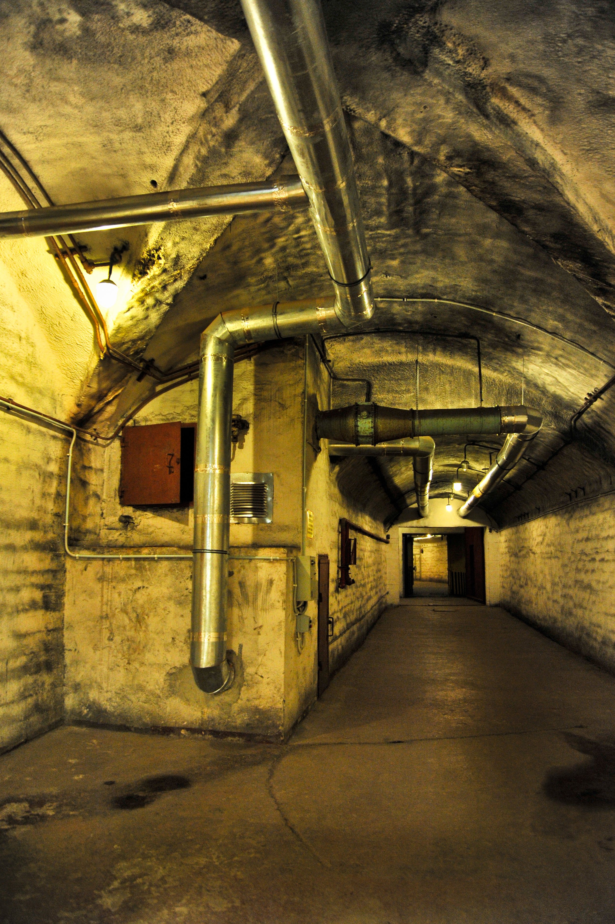 Underground tunnel with exposed metal pipes running along the ceiling and walls, dim lighting, and a concrete floor.