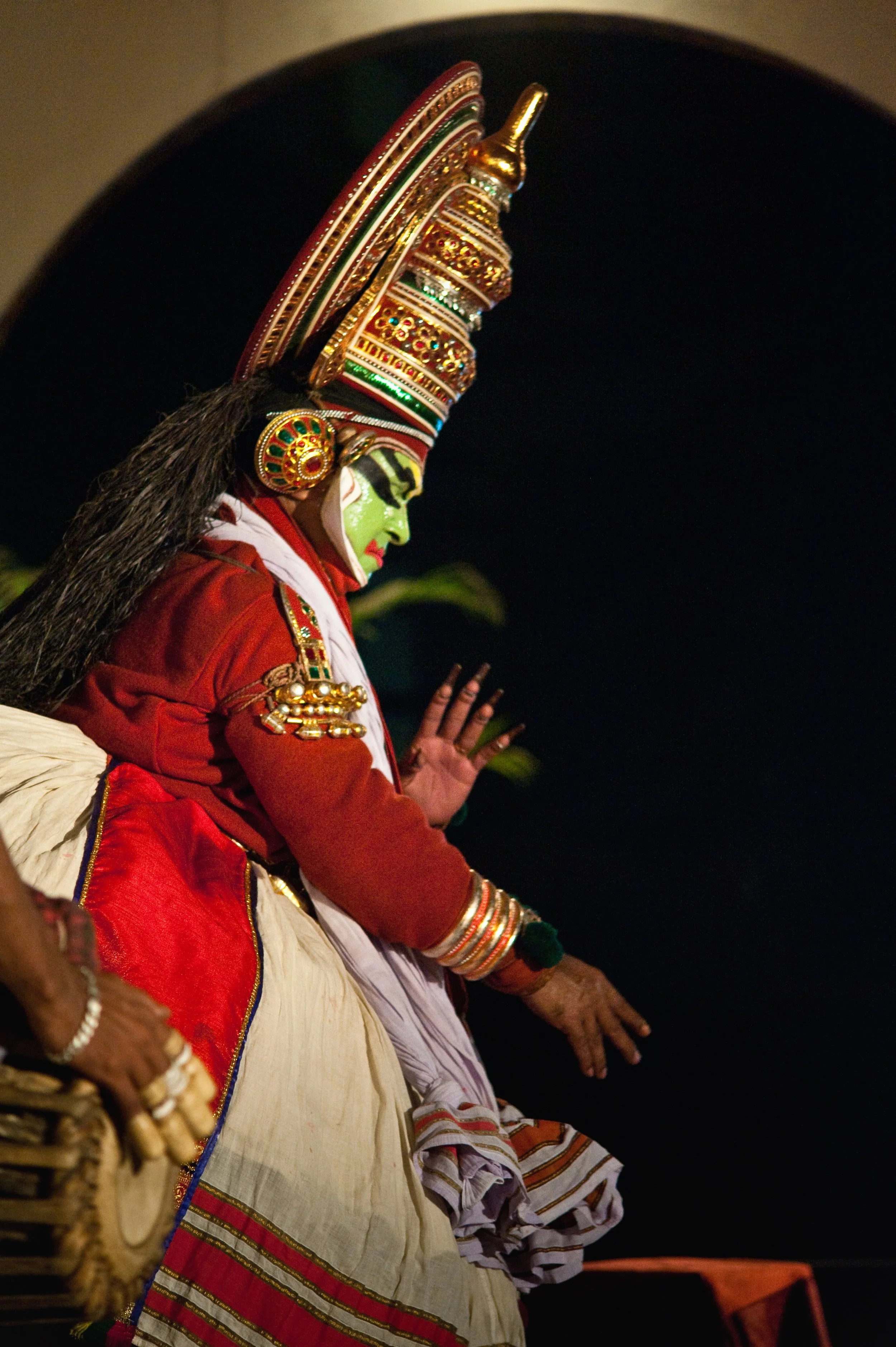 A person dressed as Lord Krishna performing a dance or ritual during a traditional celebration, wearing elaborate jewelry and a vibrant costume with face painted green, and a tall ornate crown.