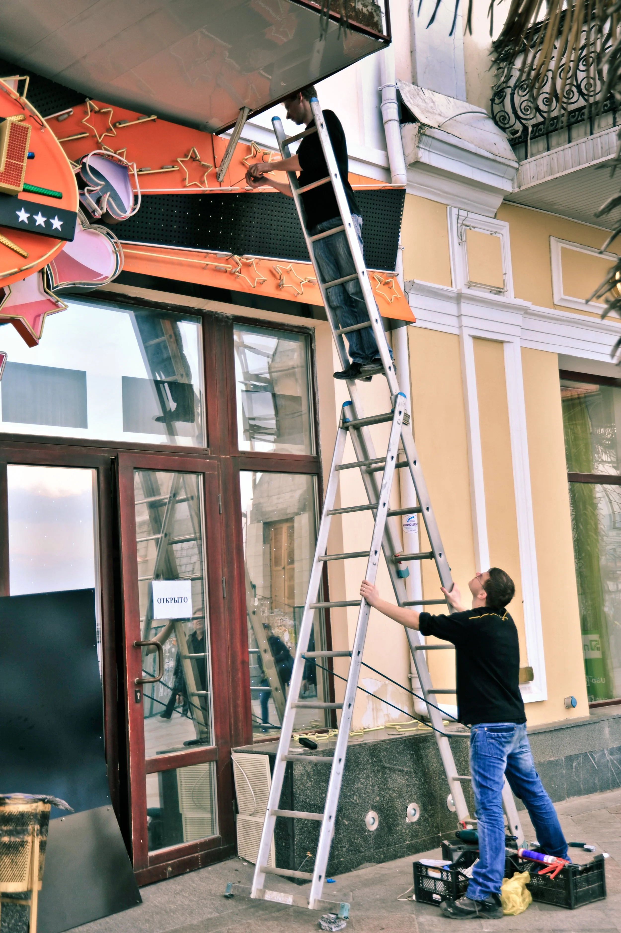Two men setting up decorative lights on a storefront exterior, one standing on an extended ladder and the other holding the ladder steady on the sidewalk.
