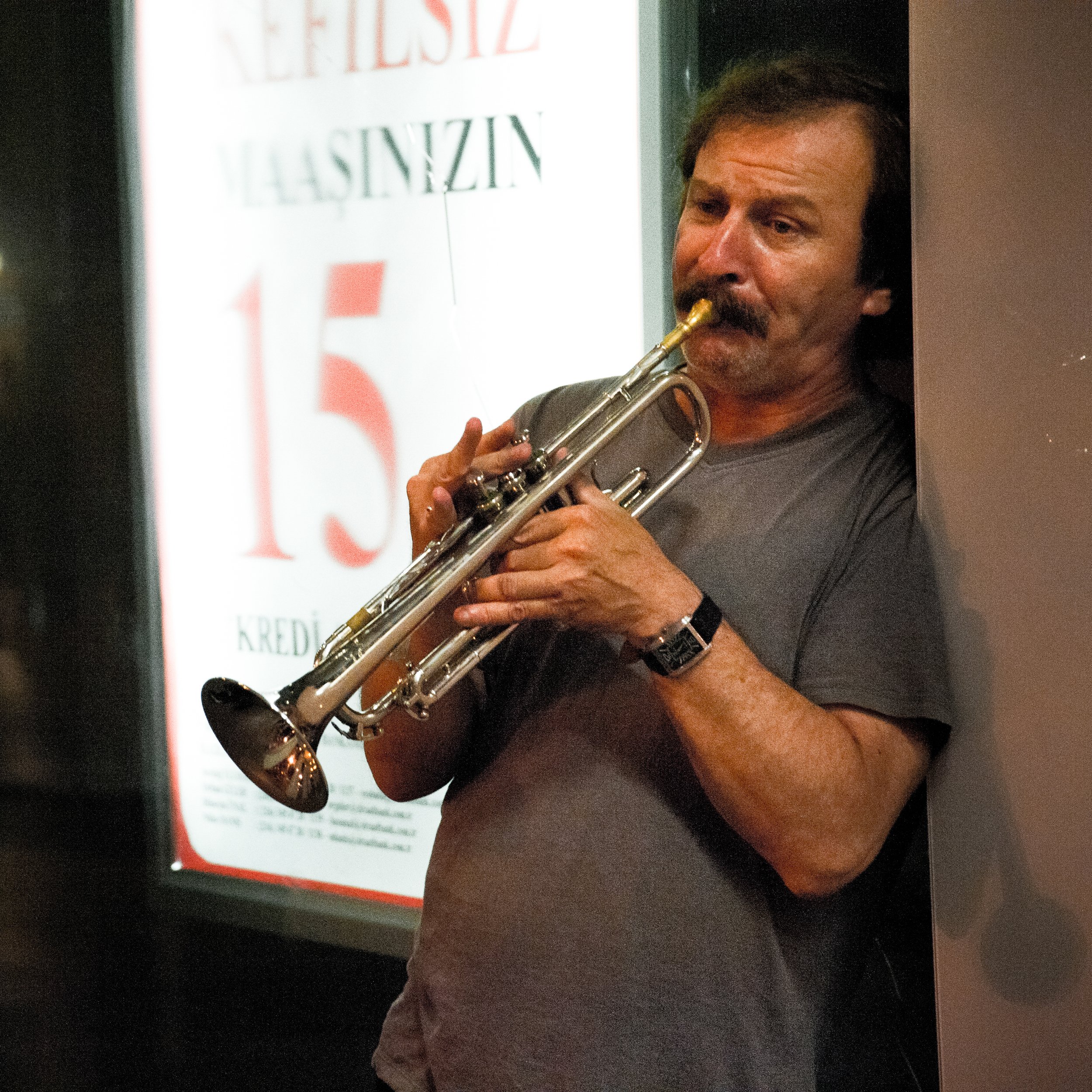 A man with dark hair, a mustache, and a beard playing a trumpet in a corner, wearing a gray t-shirt and a watch, with a sign in the background that reads verse 15.
