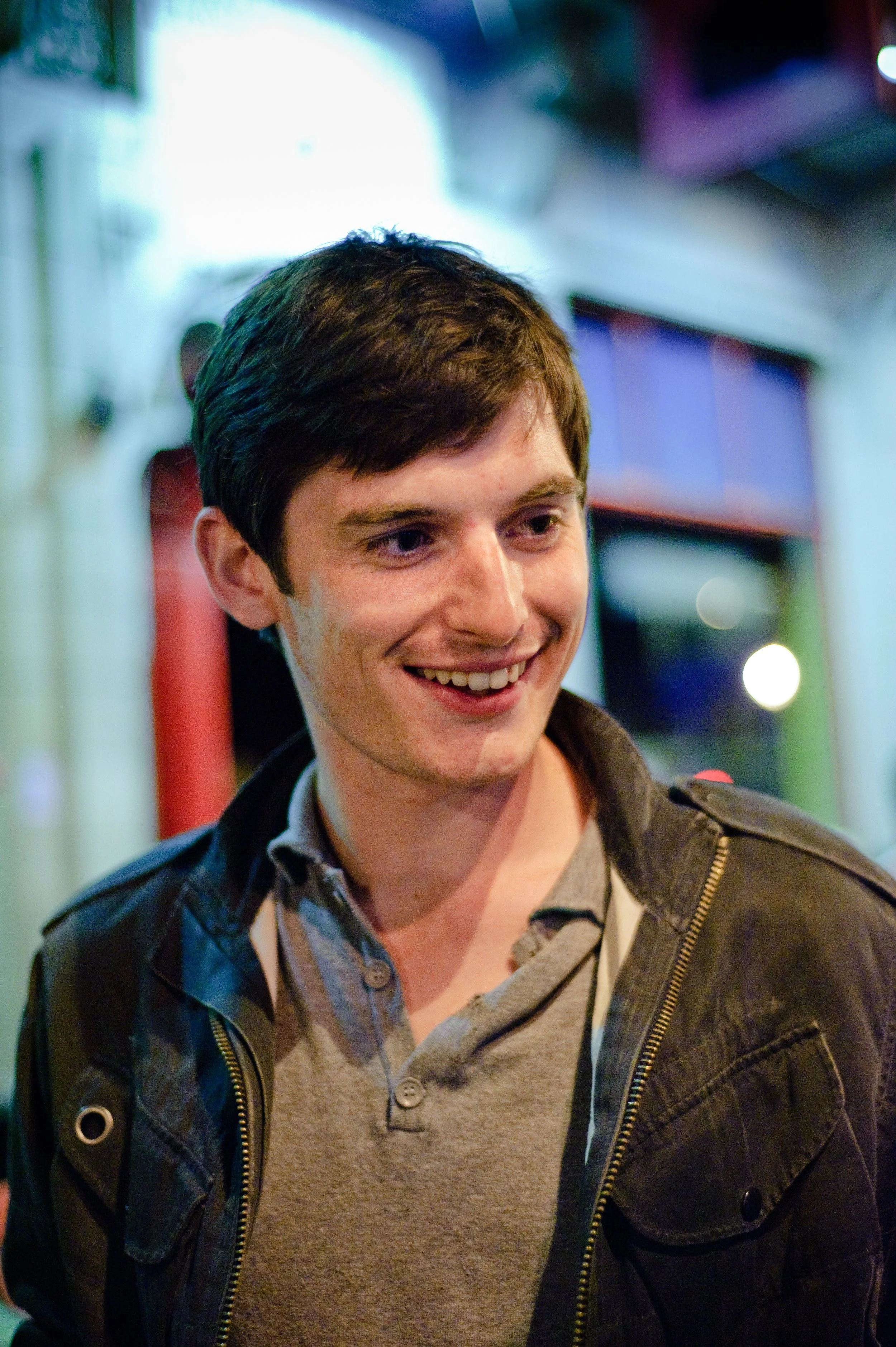 Young man with dark hair smiling, wearing a gray button-up shirt and black jacket, standing outdoors at night with colorful blurred background lights.