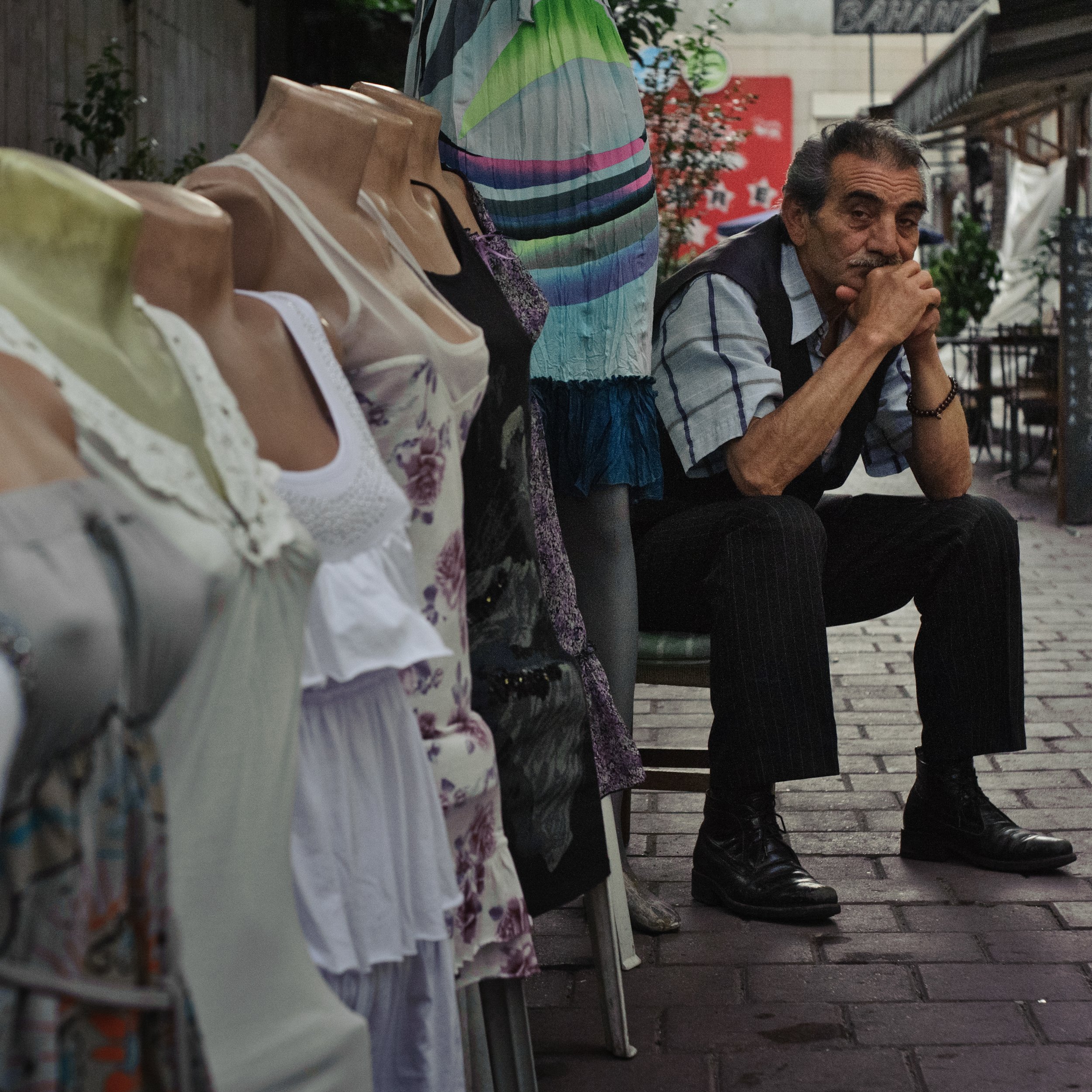 A man sitting on a bench next to a street market stall with dresses on mannequins. The man appears to be deep in thought, with hands clasped near his mouth, and is dressed in a short-sleeved plaid shirt, dark pants, and black shoes.