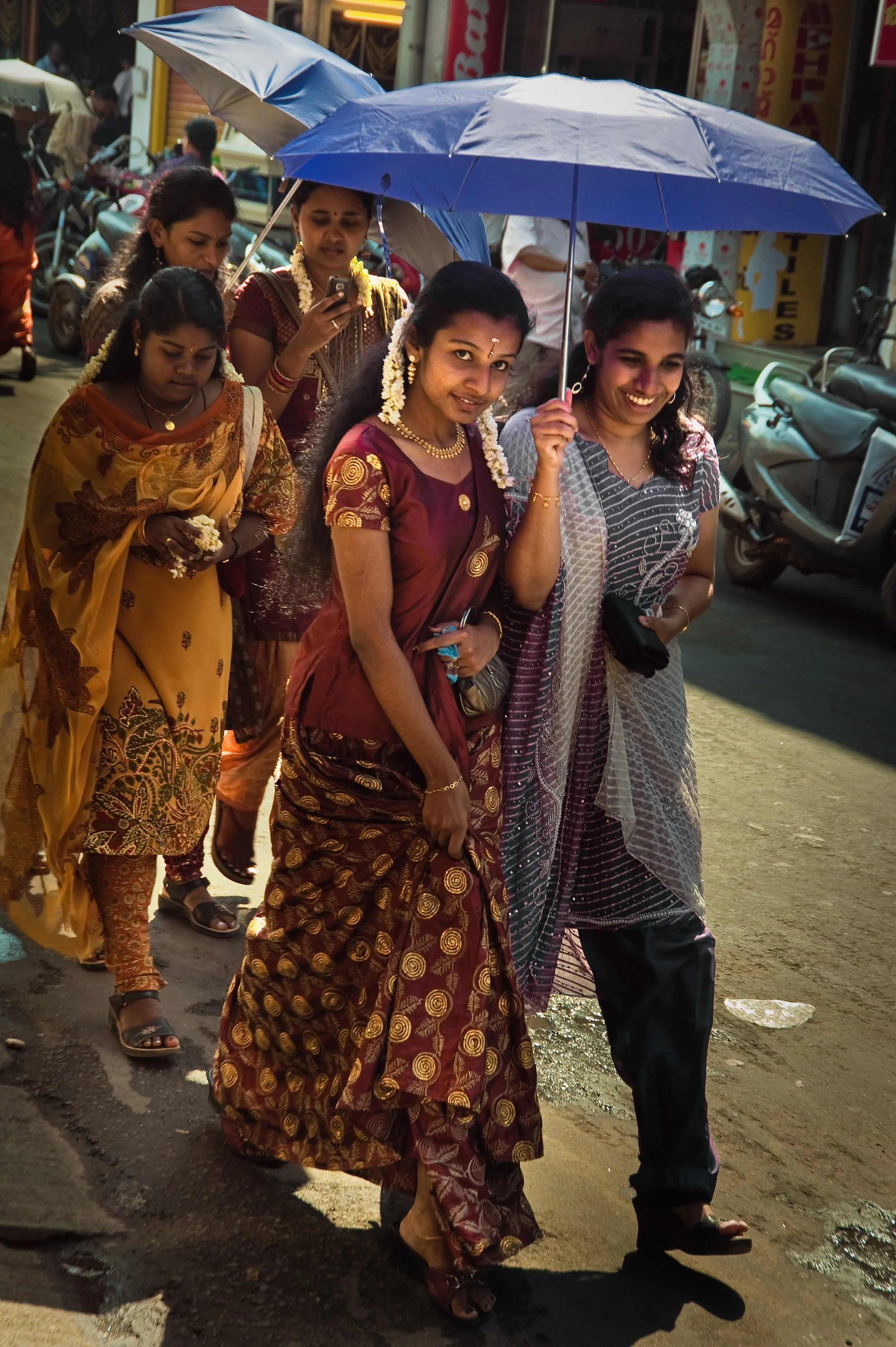 Group of women walking on a street, two women in front sharing a blue umbrella, dressed in colorful traditional Indian sarees and jewelry, some women holding flowers, with motorcycles and shops in the background.