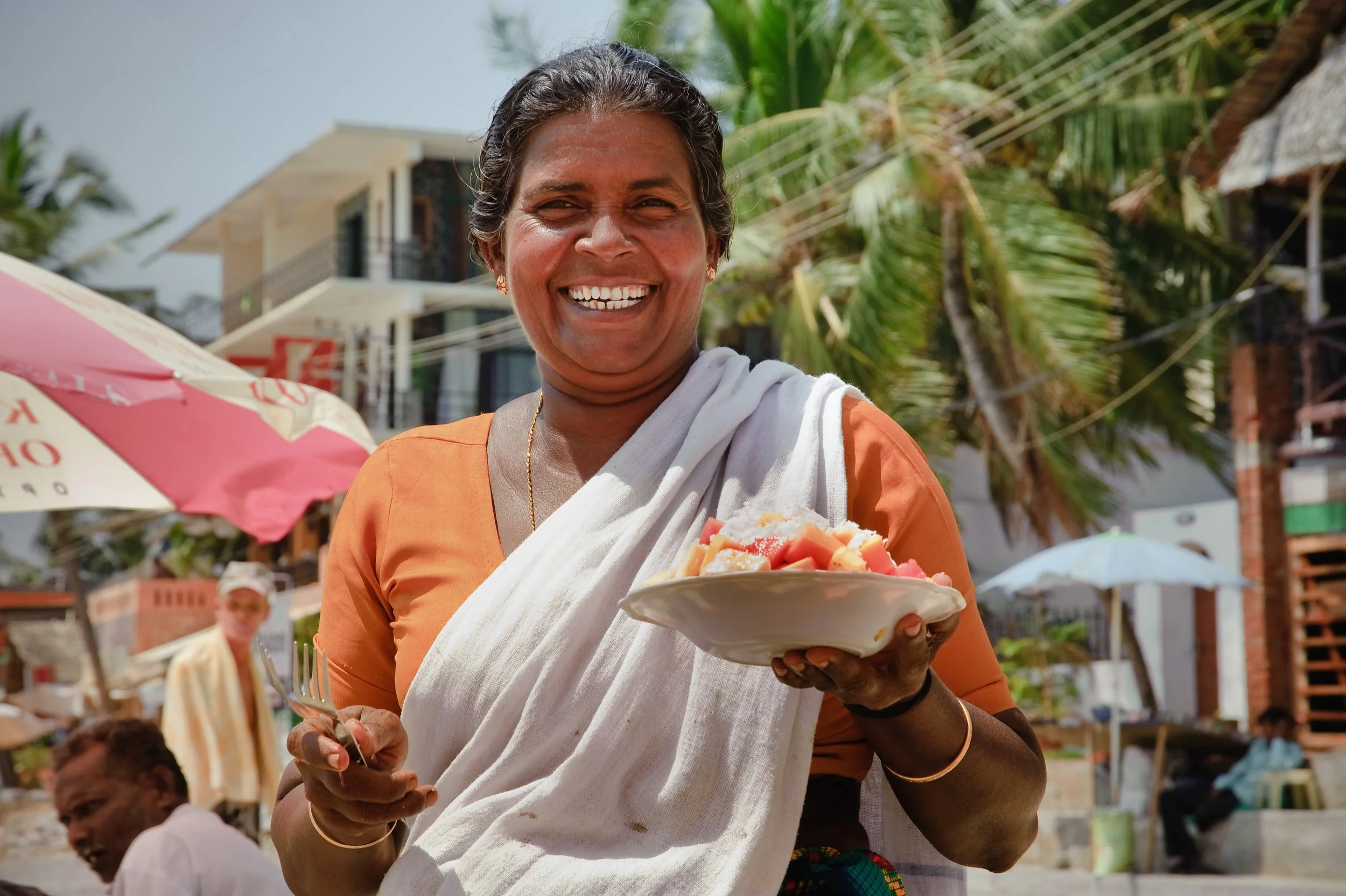 Smiling woman in orange top with white sari holding a bowl of fruit outdoors near beach umbrellas and palm trees.