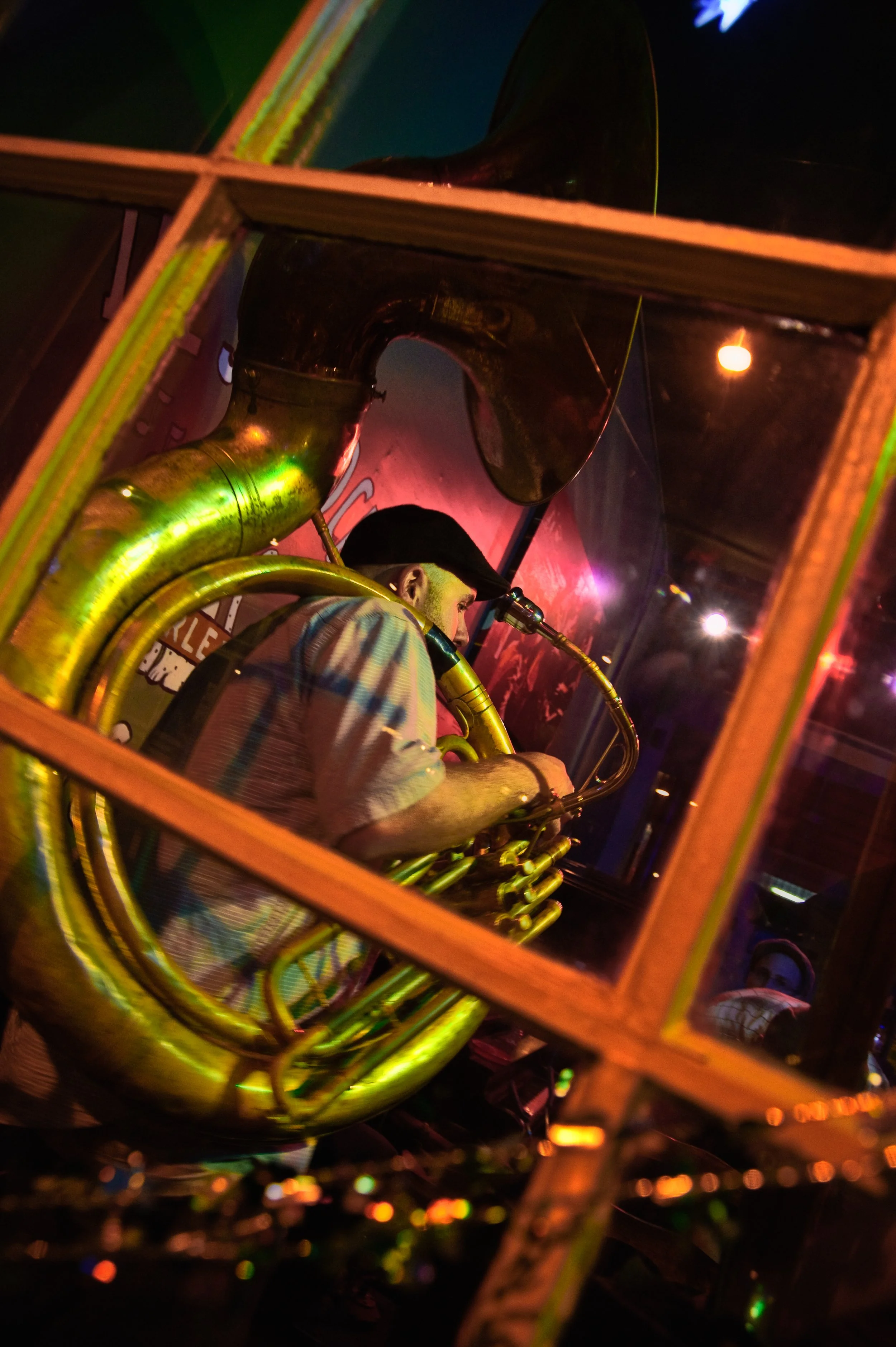 Musician playing a saxophone, photographed through a glass window, with colorful neon lighting and a reflection of the audience.