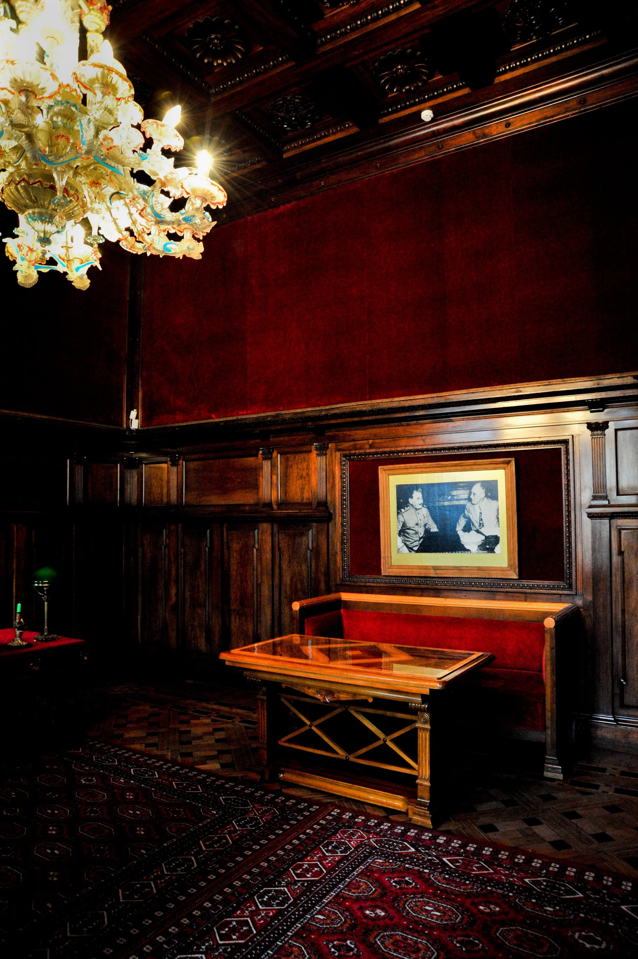 Luxurious interior of a wood-paneled room with a red velvet wall, a framed black and white photograph, a wooden table, a vintage green banker’s lamp, and a decorative chandelier.
