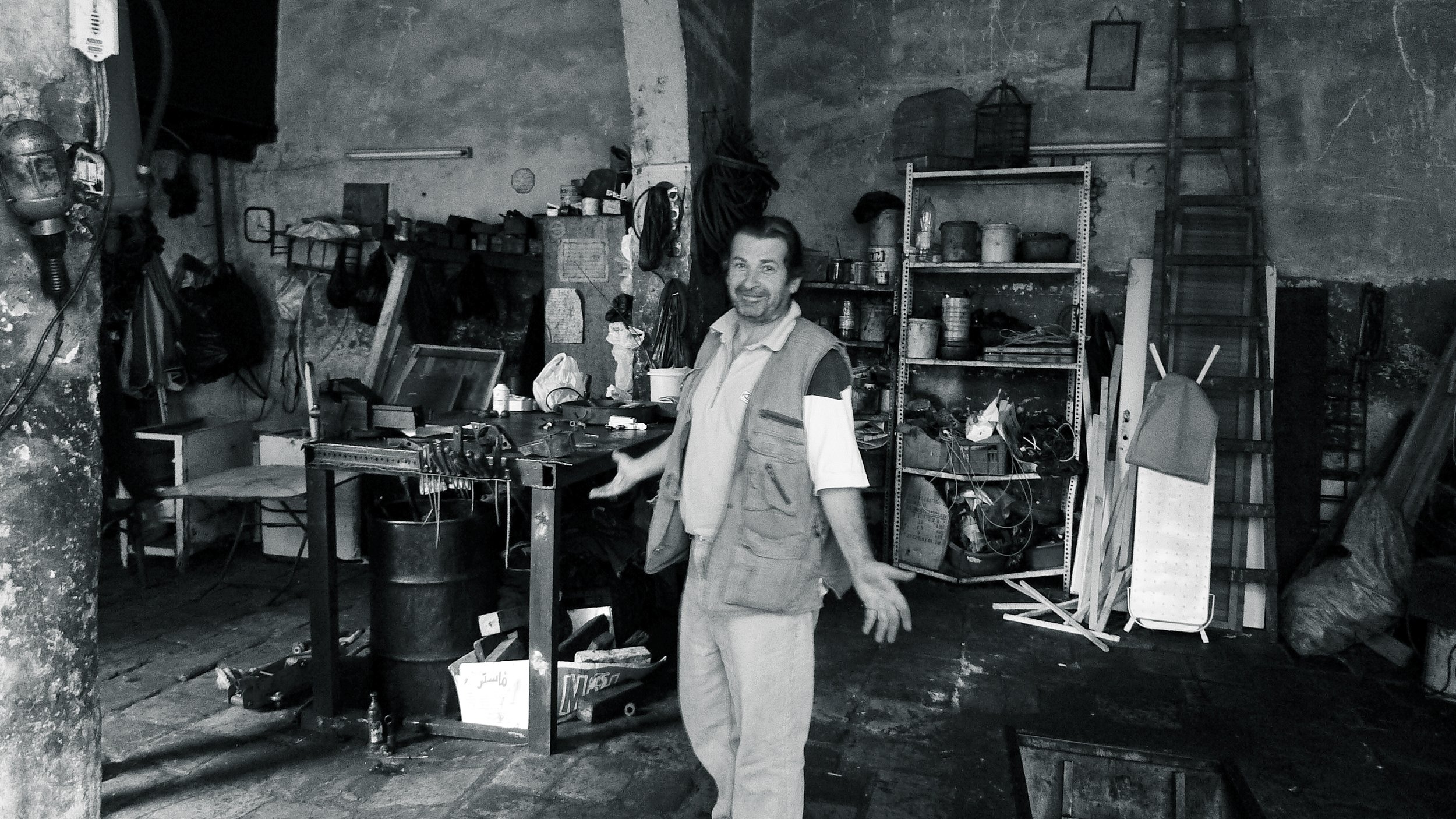 A man with a beard and white shirt standing in a cluttered workshop or garage, smiling, with tools, shelves, ladders, and other equipment around him.