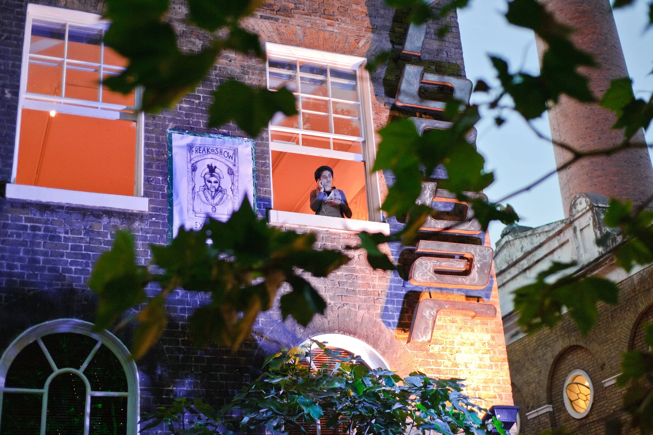 A young man standing at an open window of a brick building, looking outside. The building has a sign that reads 'FREAK SHOW' and neon lights on the wall, with leaves partially framing the scene.