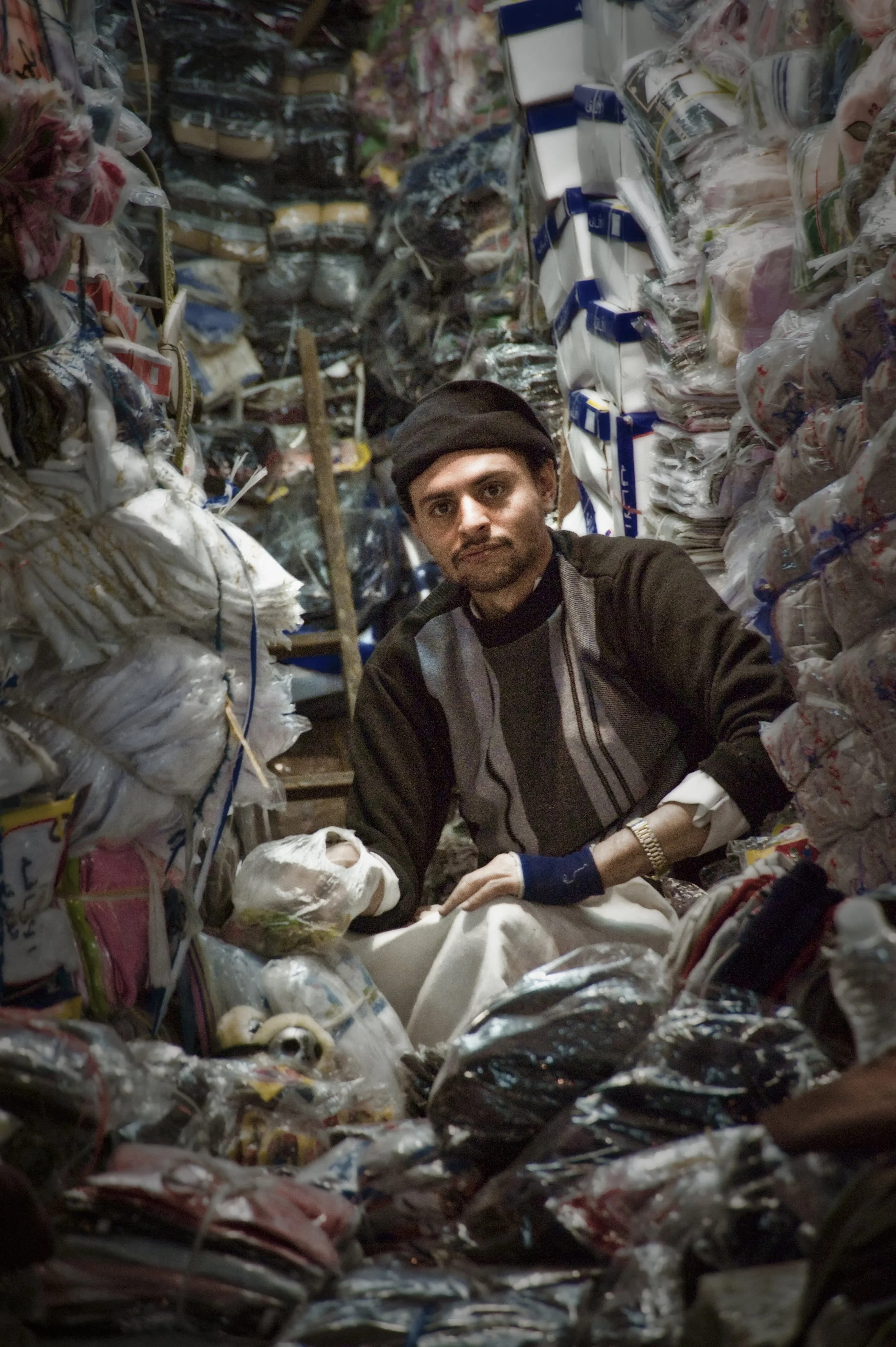 A man sitting in a narrow alley surrounded by stacked plastic bags and packaging materials in a recycling or waste collection area.
