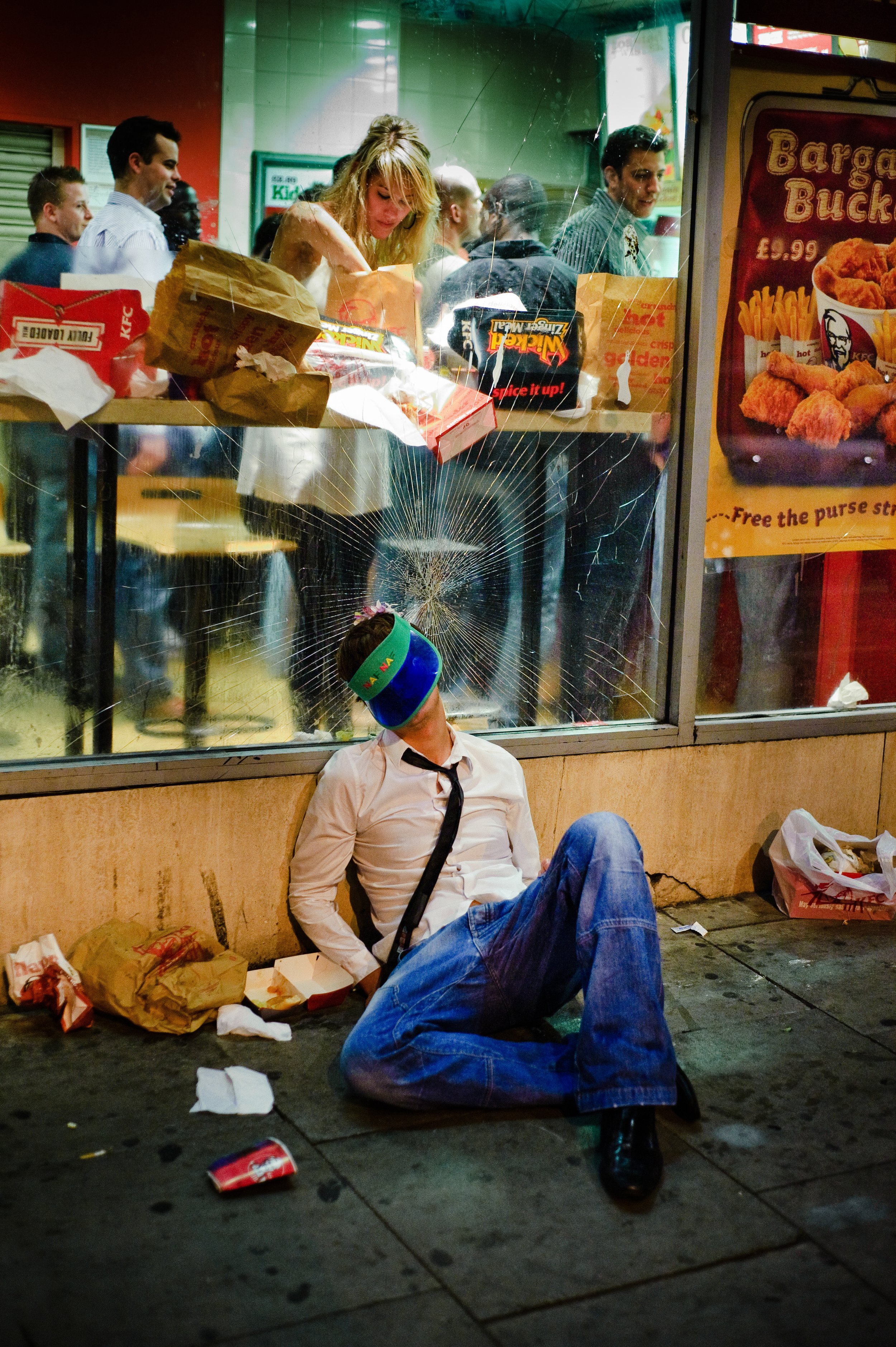 A man wearing a white shirt, black tie, and jeans is sitting on the sidewalk with his head leaning against a window with a crack in the glass. He has a colorful visor over his eyes and is surrounded by fast food wrappers and debris. Inside the fast f