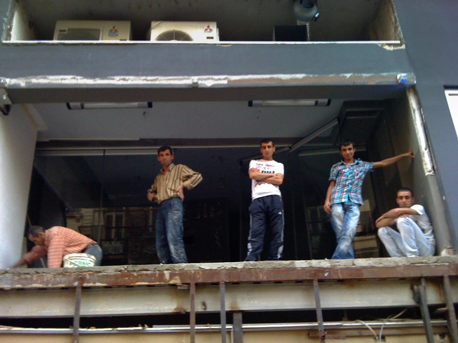 Five young men on an unfinished building balcony; one is sitting on the floor, two are standing with crossed arms, one has his hand on the wall, and the last one is standing with his hand on his hip.