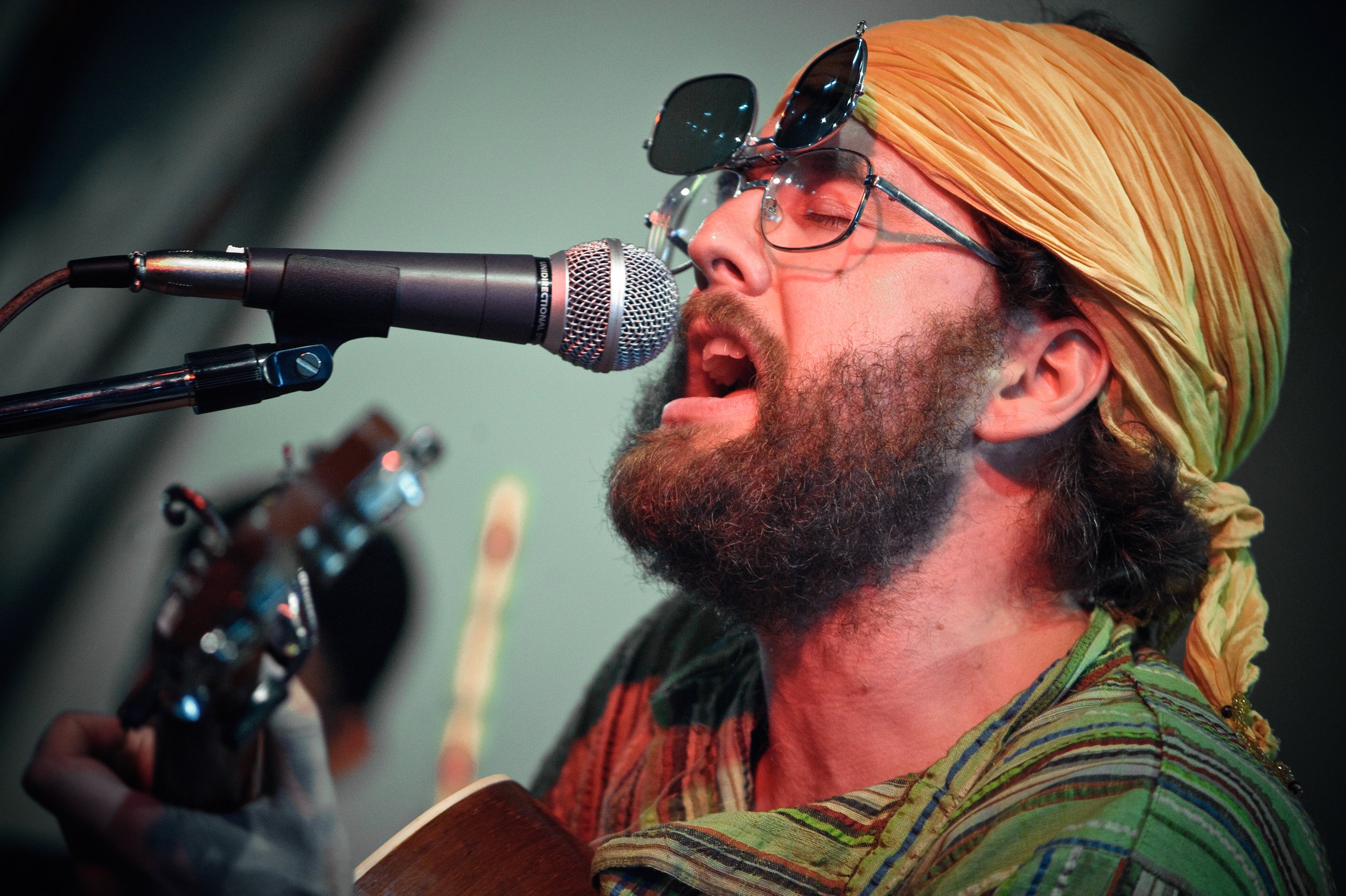 A man singing into a microphone while playing a guitar, wearing glasses, a yellow headscarf, and a colorful shirt.